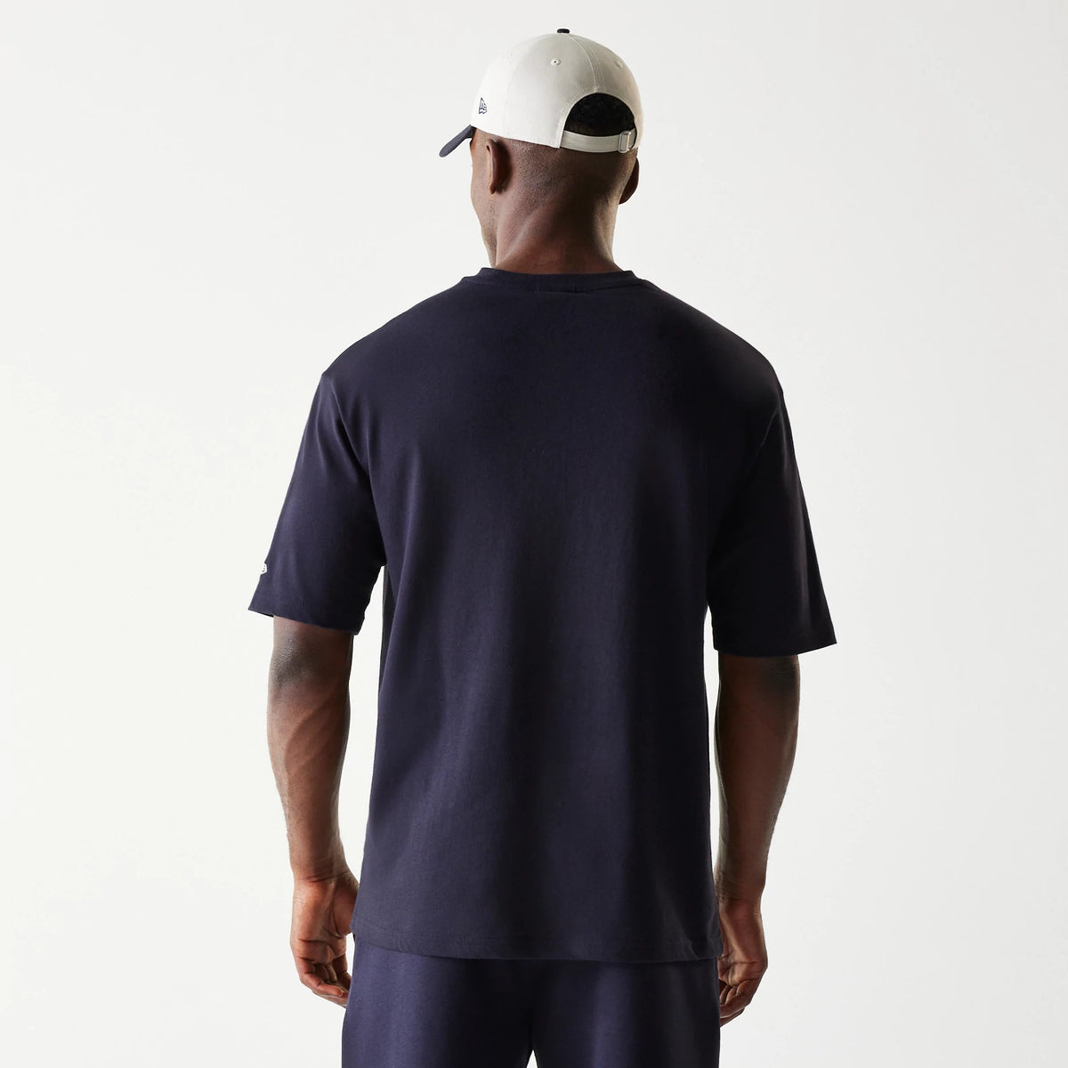 New Era Navy Oversized T-Shirt, back view on a model showing the plain navy back with short sleeves, styled with a white cap featuring a small embroidered New Era logo on the side.