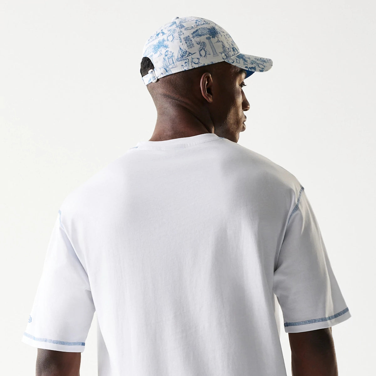 New Era Orthodox Vase White Oversized T-Shirt, back view on a model also wearing a patterned cap, highlighting the plain white back with blue stitching on the sleeves and an embroidered New Era logo on the left sleeve.
