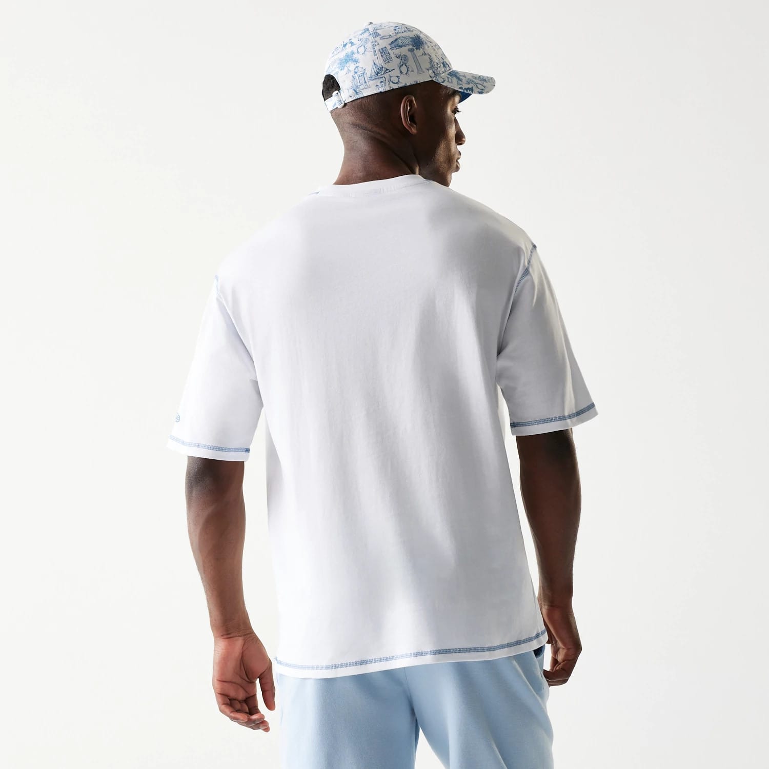 New Era Orthodox Vase White Oversized T-Shirt, back view on a model wearing a blue patterned cap, showing clean white fabric with contrasting blue stitching along the hem and sleeves, with a embroidered New Era logo on the sleeve.