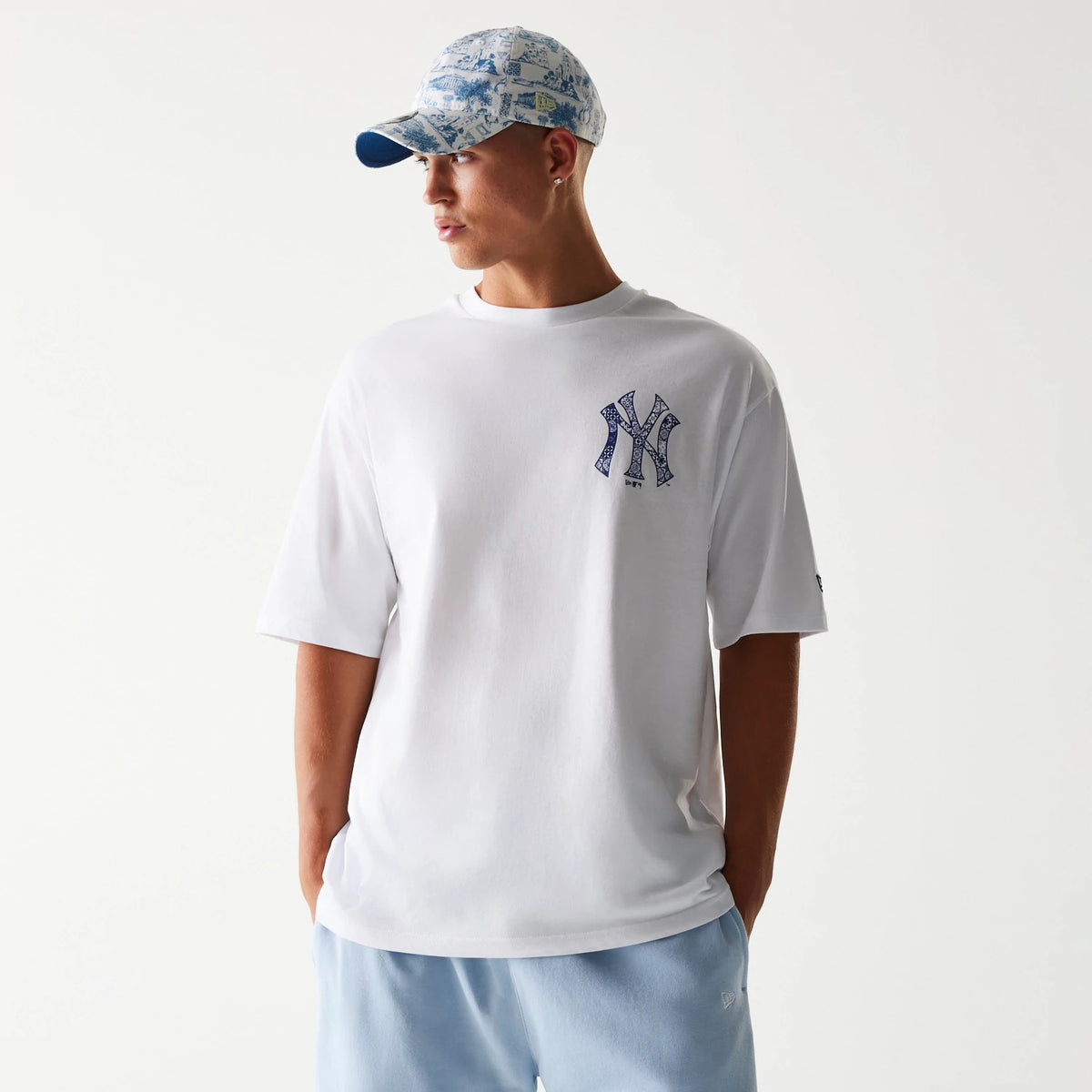 New Era New York Yankees MLB White Oversized T-Shirt, front view on a model styled with a patterned cap and light blue trousers, featuring a large embroidered Yankees logo in blue on the left chest and a New Era flag on the sleeve.