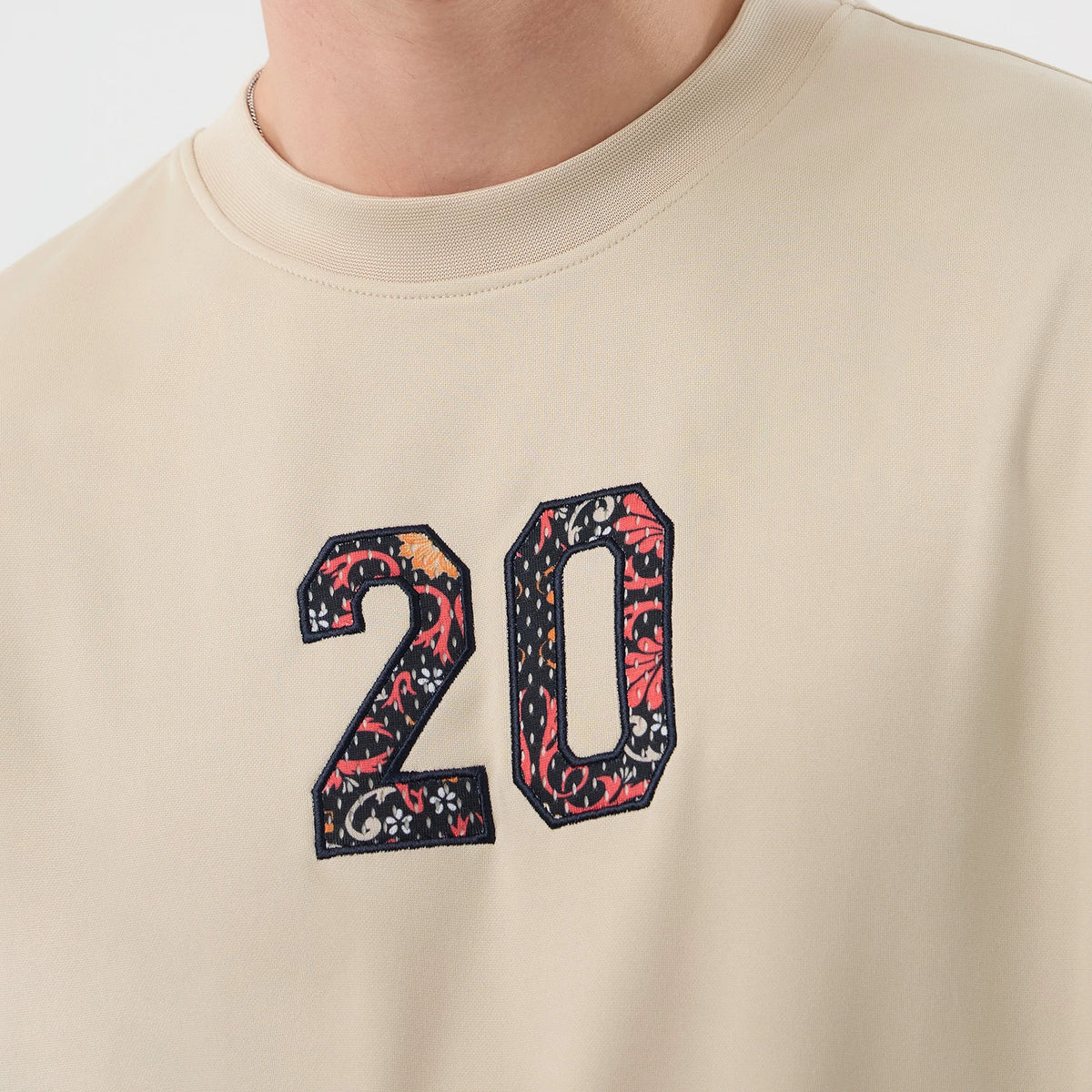 New Era Mesh Infill Light Beige Oversized T-Shirt, front view showing the intricately embroidered number 20 in colourful patterns centred on the light beige fabric near the neckline.
