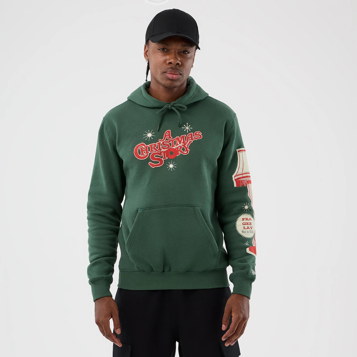 New Era A Christmas Story Major Award Dark Green Pullover Hoodie, front view on model with a black cap, showing red and white lettering on the chest, a unique graphic on the right sleeve, and an embroidered New Era logo near the cuff.