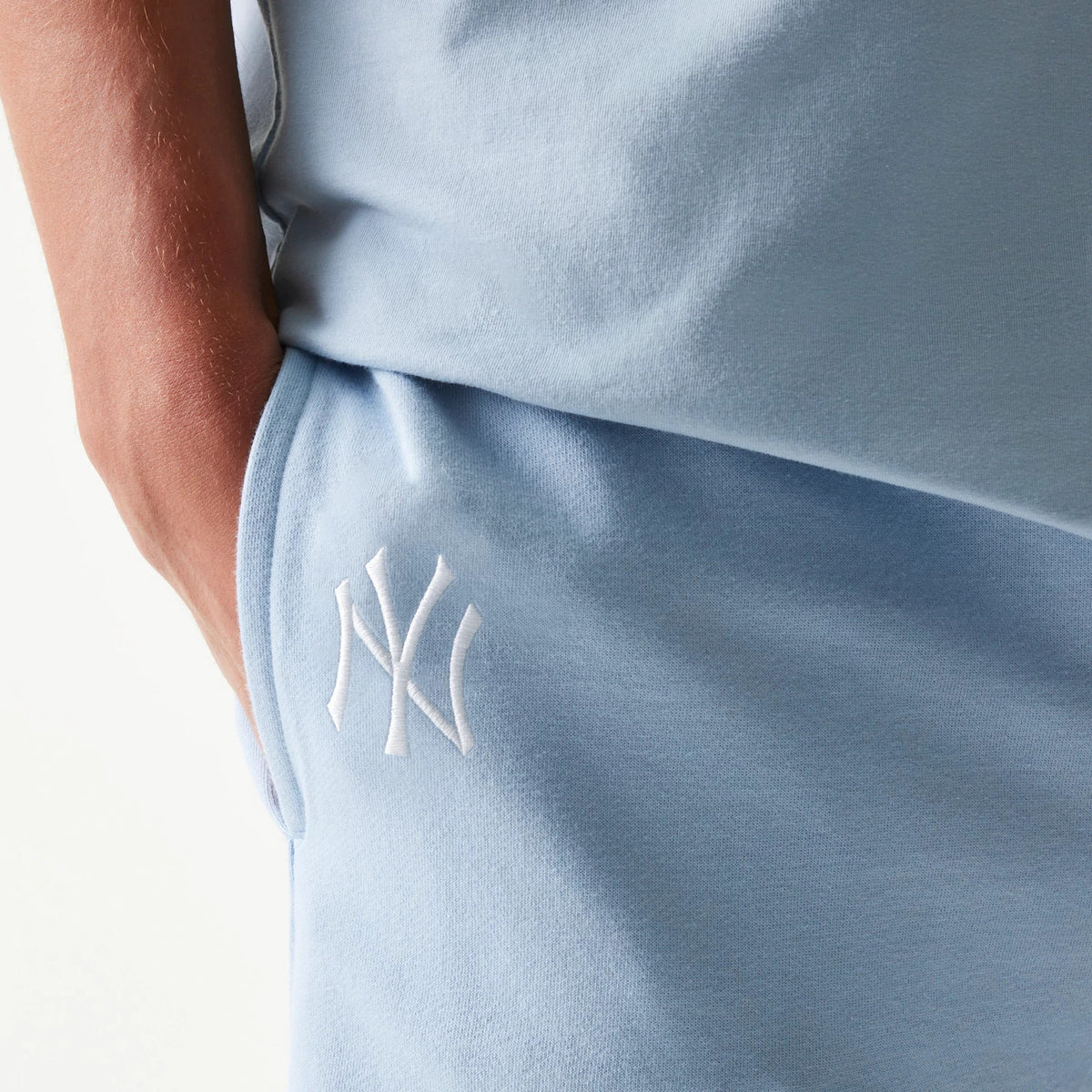 New Era New York Yankees MLB League Essential Midi Pastel Blue Shorts, close-up front view showing the light blue fabric with an embroidered white New York Yankees logo near the pocket on the left thigh.