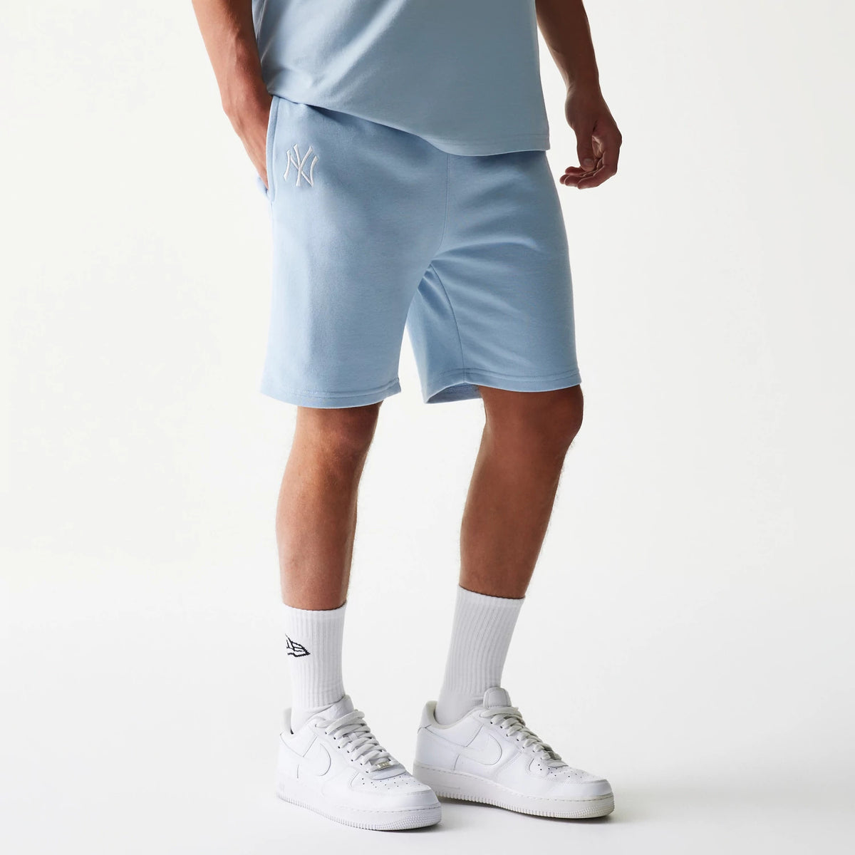 New Era New York Yankees MLB League Essential Midi Pastel Blue Shorts, front three-quarter view on a model showing the pastel blue fabric with an embroidered white New York Yankees logo on the left leg, paired with white trainers and socks.