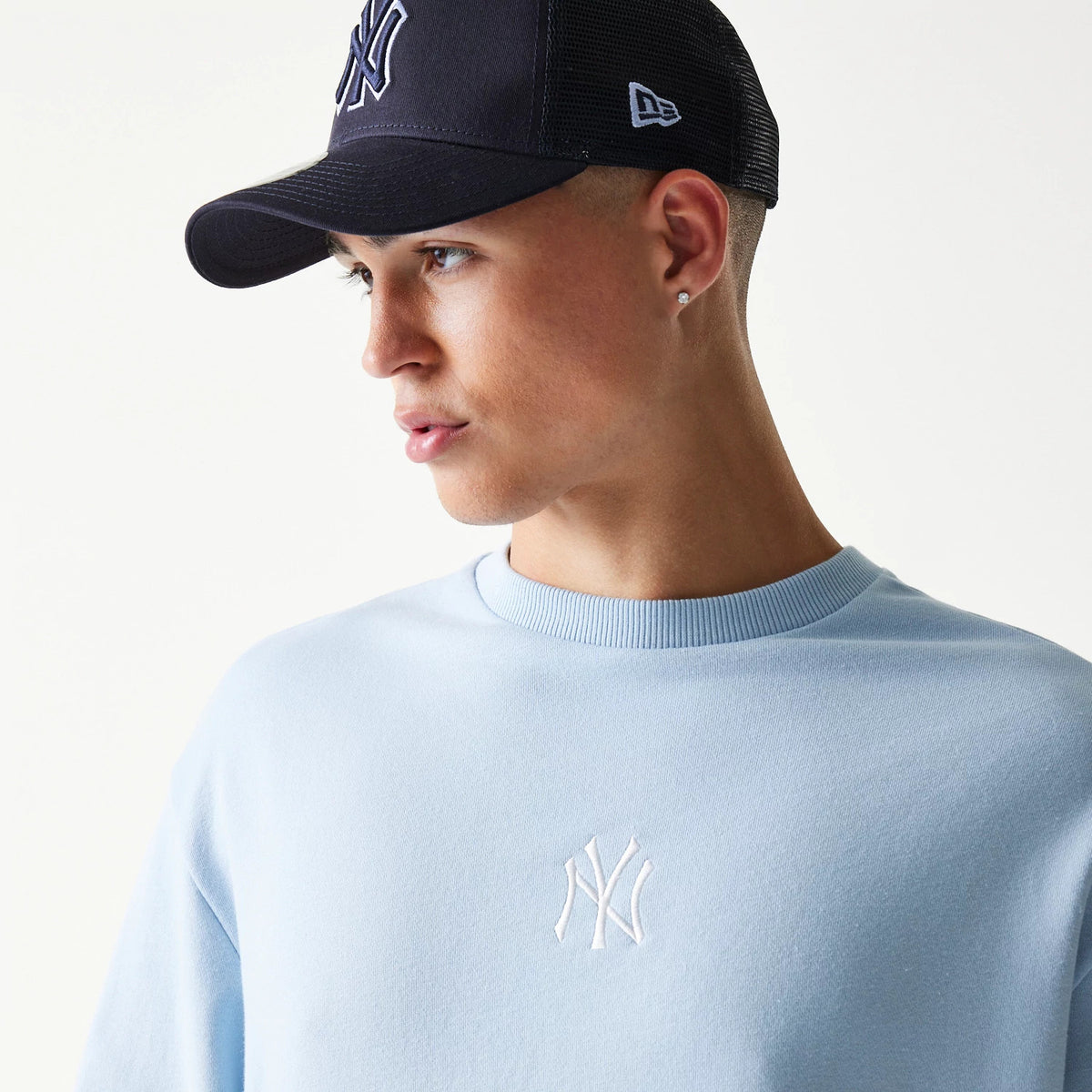 New Era New York Yankees MLB Midi Blue Oversized Crew Neck Sweater, on-model view featuring the white embroidered NY logo on the chest with a light blue ribbed collar, paired with a black cap showing the embroidered NY logo on the front.