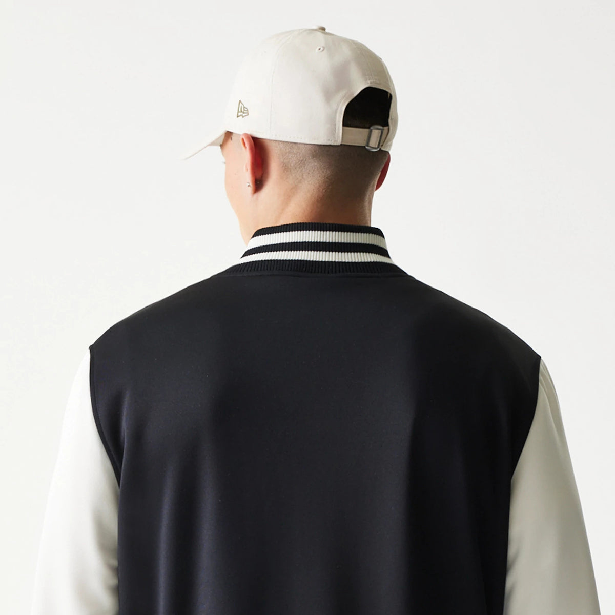 New Era Chicago Bulls NBA Black Bomber Jacket, back view on-model featuring a solid black back with white sleeves and a contrasting black and white striped collar, styled with a white cap with an embroidered New Era logo on the side.