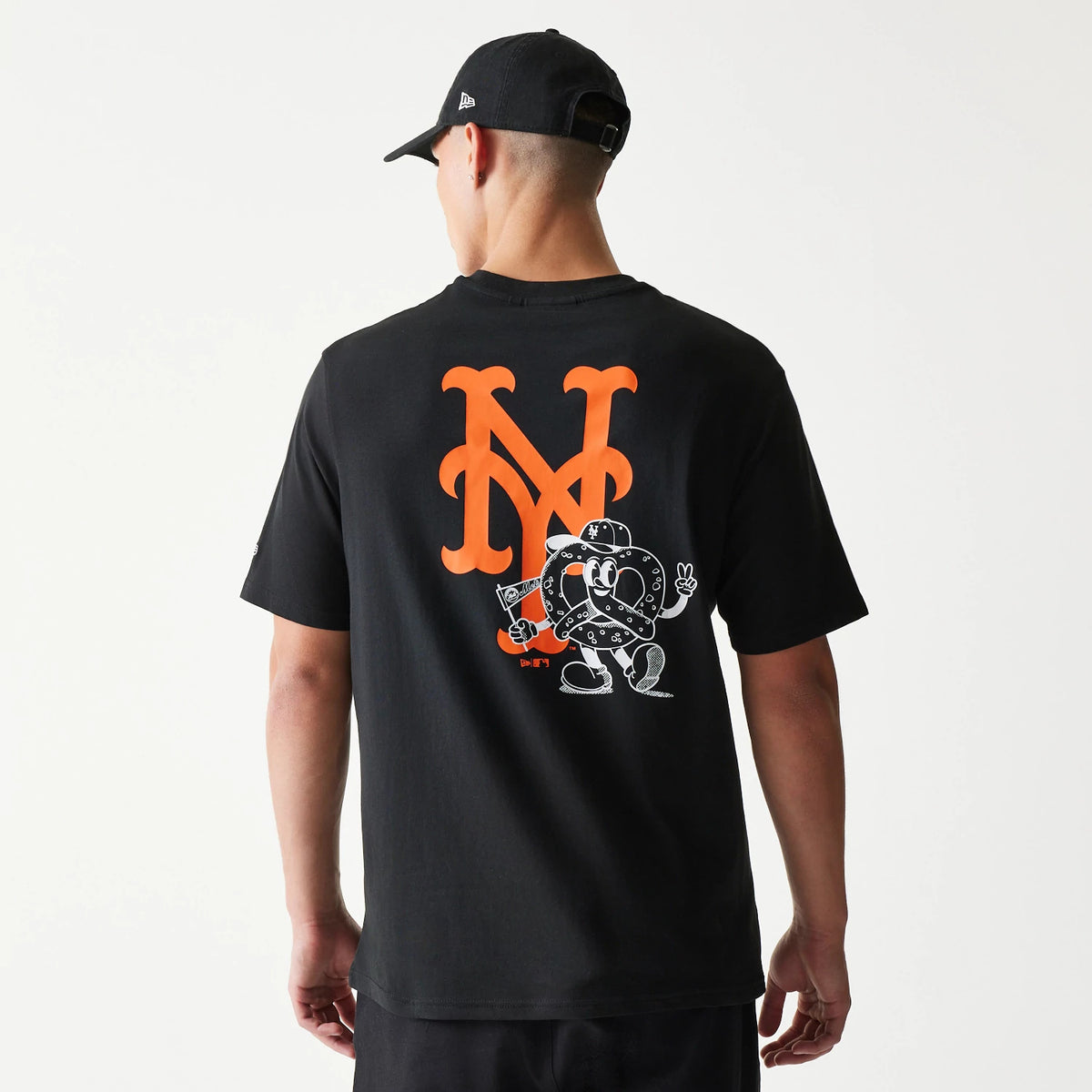 New Era New York Mets MLB Food Graphic Black T-Shirt, back view on-model featuring a large orange NY logo and a cartoon pretzel character graphic on the lower section, styled casually with a black cap.