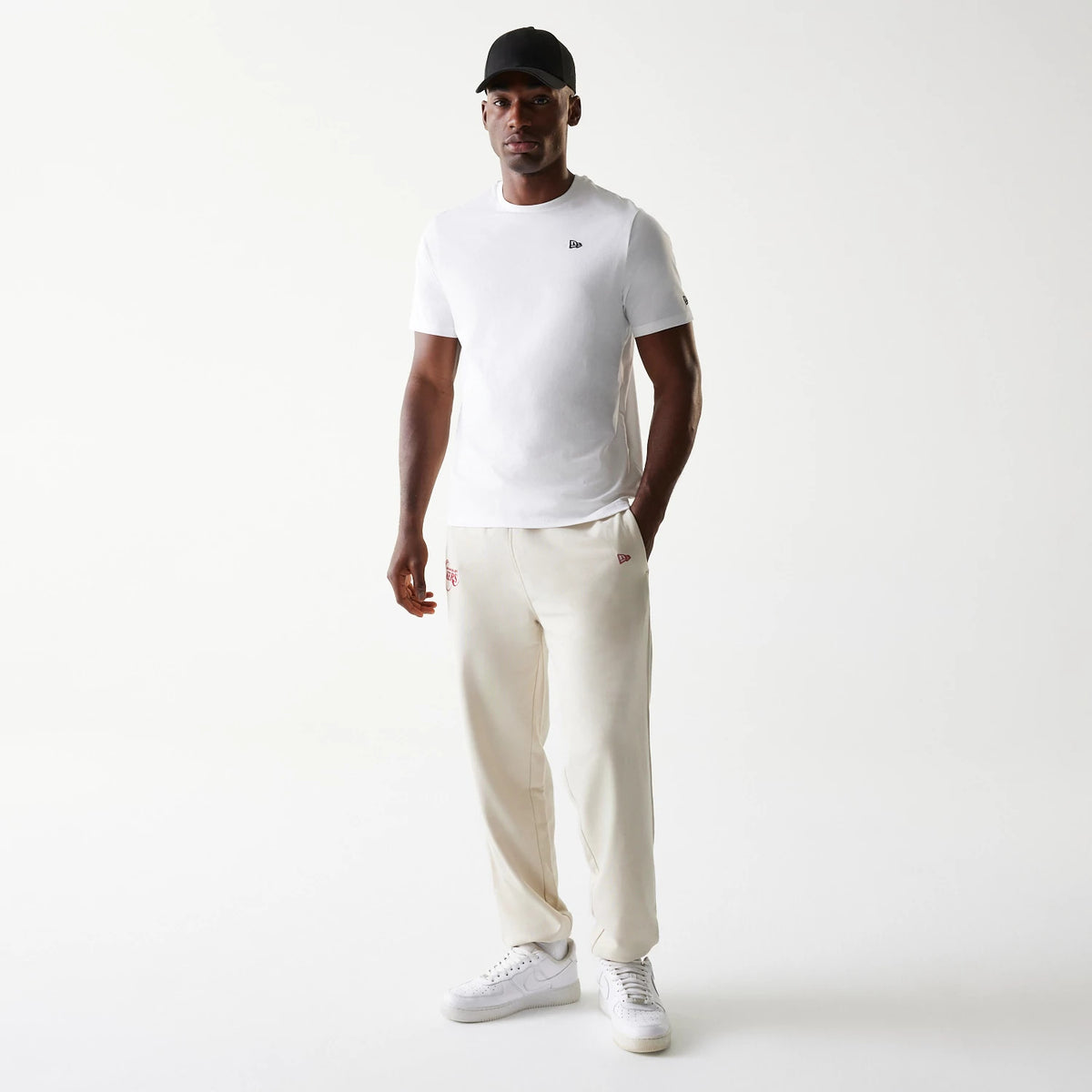 New Era LA Lakers NBA League Essential Cream Joggers, on-model view showing cream joggers with red LA Lakers logo on the right thigh, New Era flag on the left thigh, paired with a white T-shirt and white trainers.