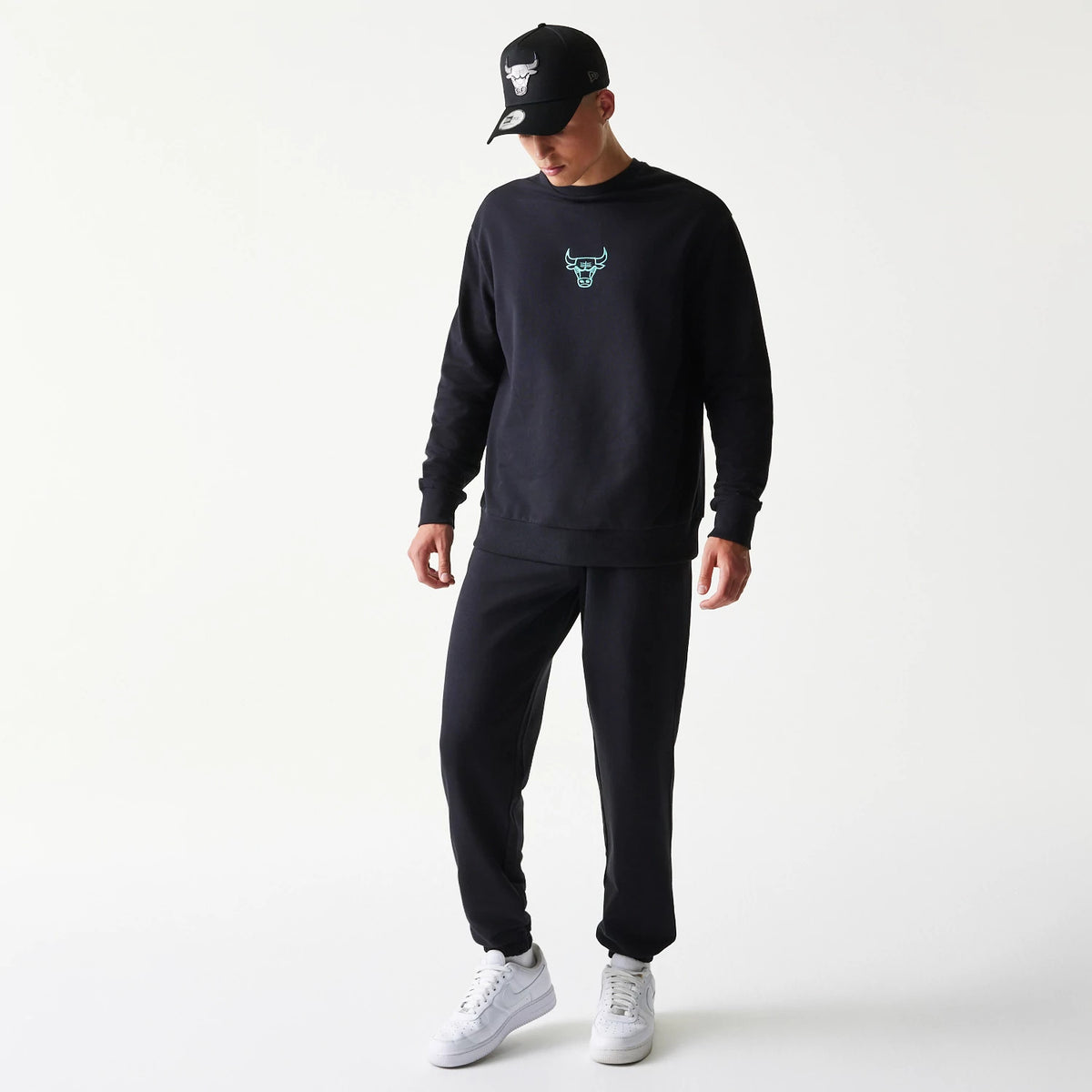 New Era Chicago Bulls NBA League Essential Black Joggers, on a model with matching black sweatshirt and cap, featuring a relaxed fit and elasticated cuffs, styled with white trainers for a casual look.