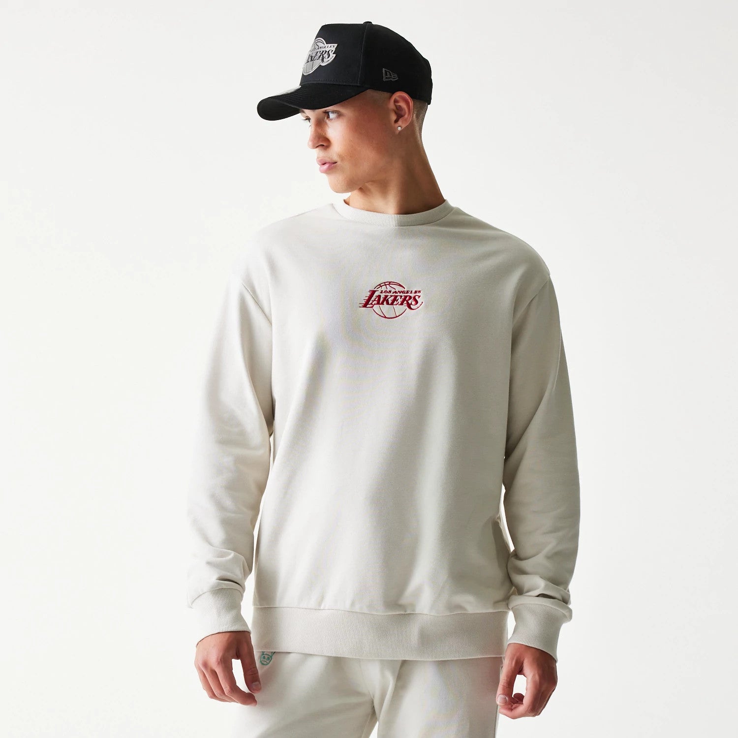 New Era LA Lakers NBA Cream Oversized Crew Neck Sweater, front view on a model wearing a black cap, showing the red embroidered Lakers logo centred on the chest and ribbed cuffs and hem.