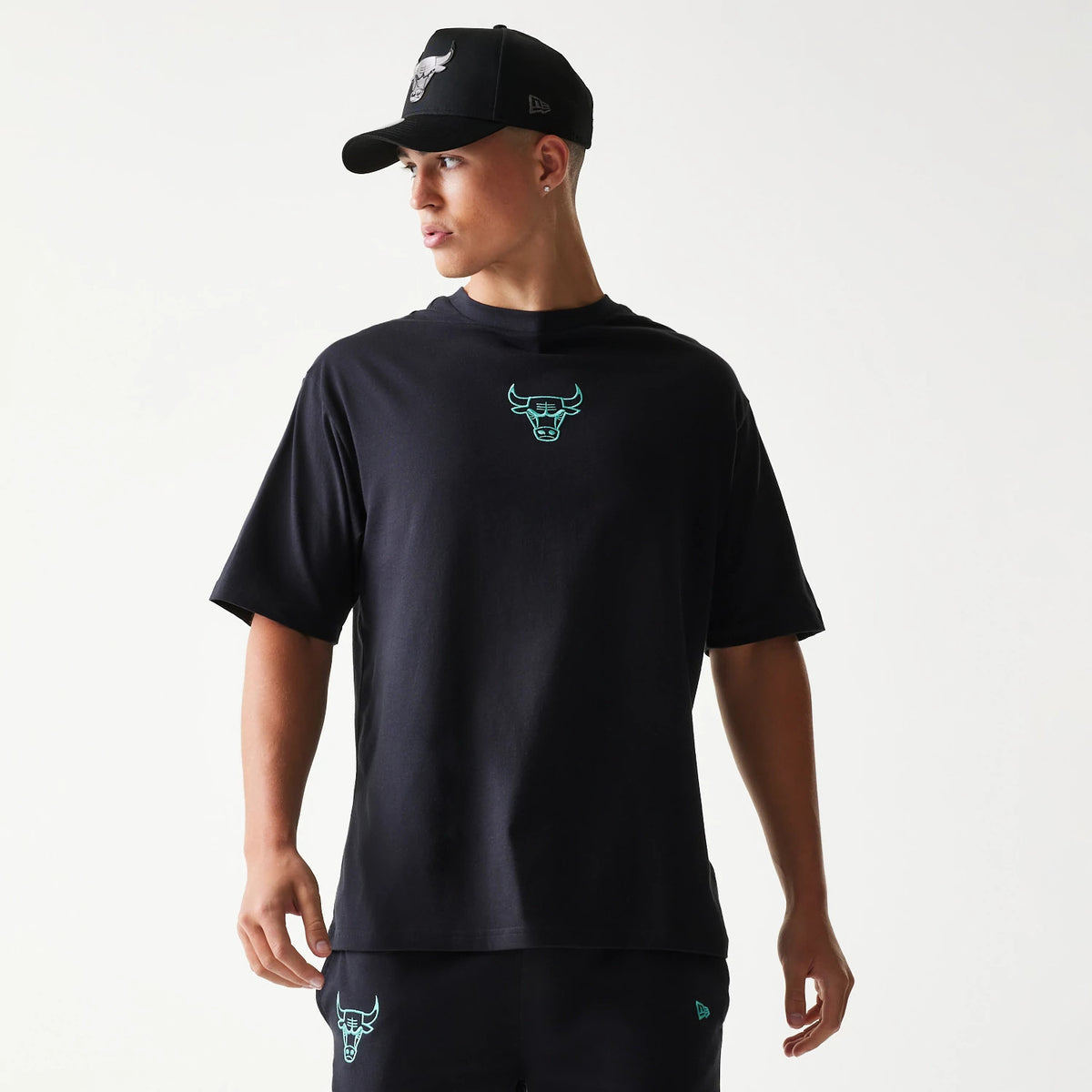 New Era Chicago Bulls NBA League Essential Black Oversized T-Shirt, front view on a model with matching shorts and cap, showing an aqua bull graphic centred on the chest and an embroidered New Era logo on the left sleeve.