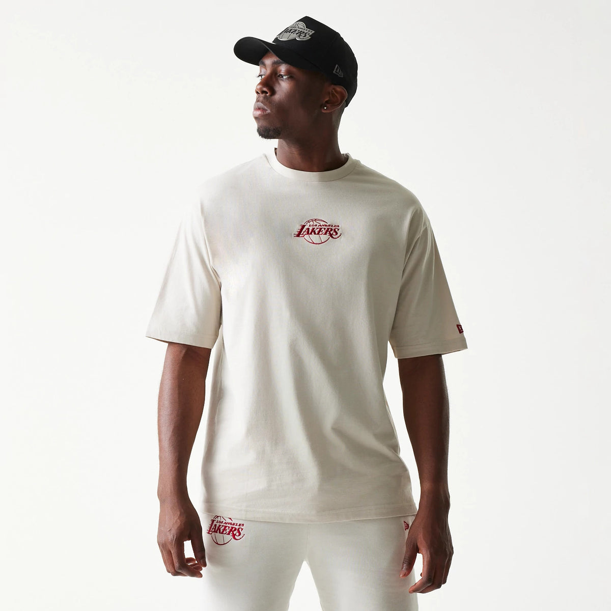 New Era LA Lakers NBA League Essential Cream Oversized T-Shirt, front view on-model showing the red embroidered Lakers logo centred on the chest, with short sleeves and a relaxed fit, styled with matching cream trousers and a black cap.
