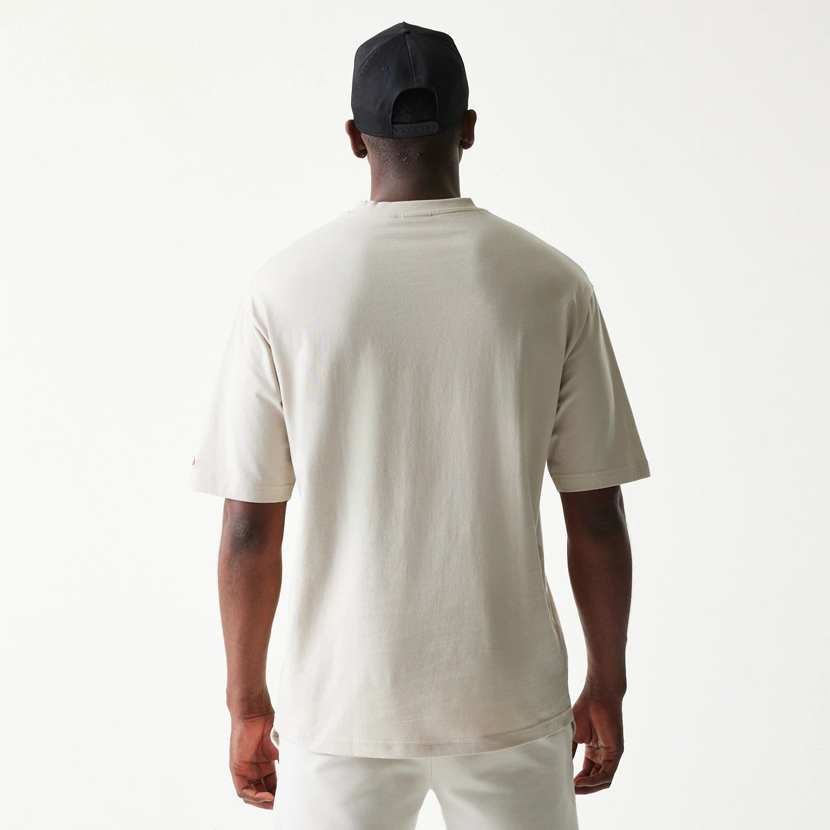 New Era LA Lakers NBA League Essential Cream Oversized T-Shirt, back view on a model styled with a black cap, showing the plain cream back and loose short sleeves, highlighting the oversized fit of the top.