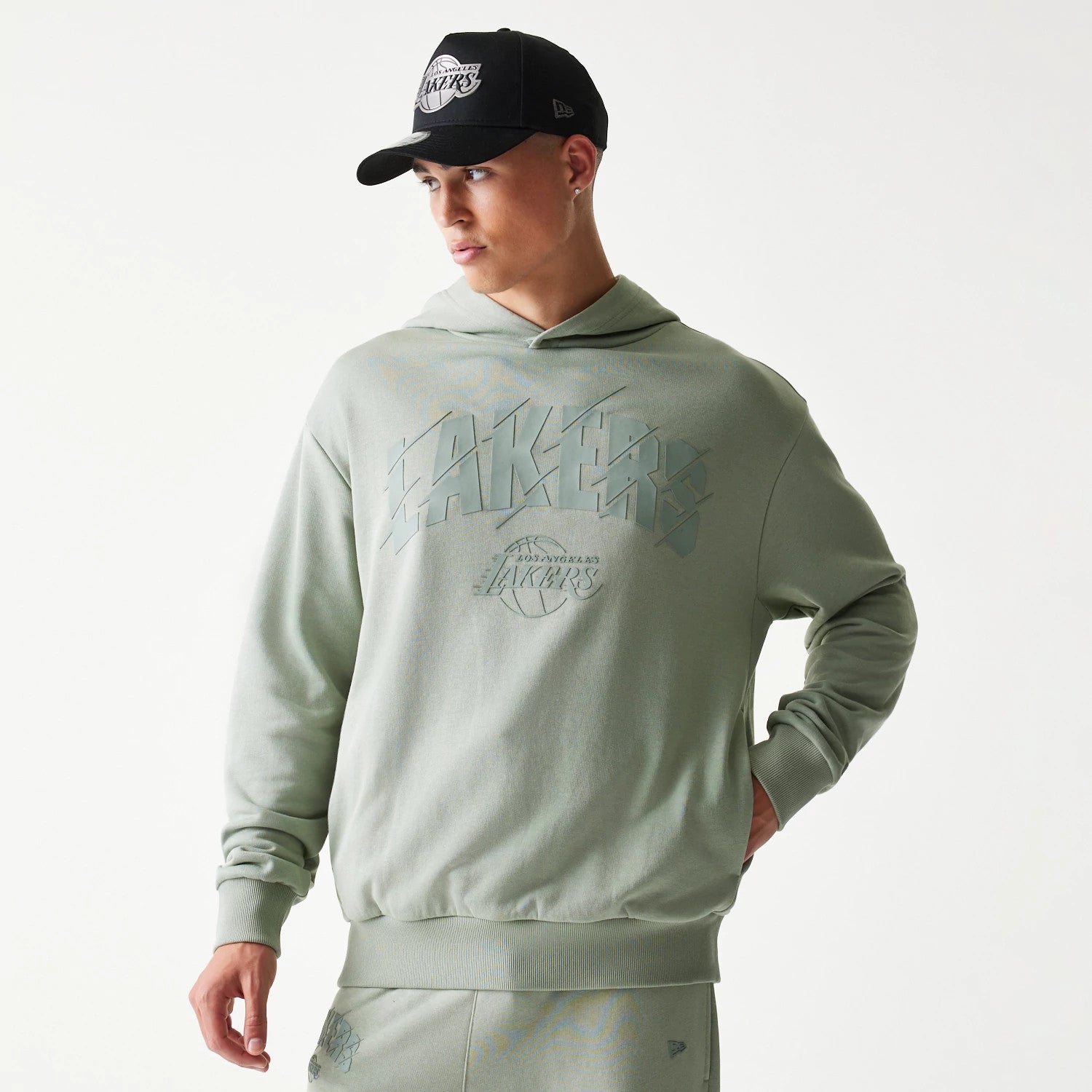 New Era LA Lakers NBA Pastel Green Oversized Pullover Hoodie, front view on a model with a black cap, featuring a large pastel green LAKERS graphic across the chest with a debossed New Era logo near the waistband.