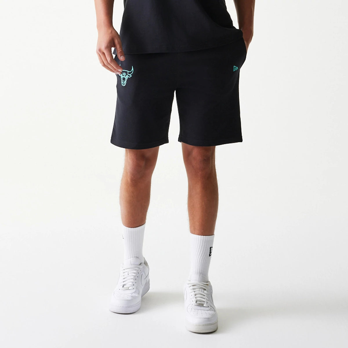 New Era Chicago Bulls NBA League Essential Black Shorts, front view on-model showing the turquoise embroidered Bulls logo near the left hem and the New Era flag embroidered above the right hem, paired with casual white trainers and tall socks.