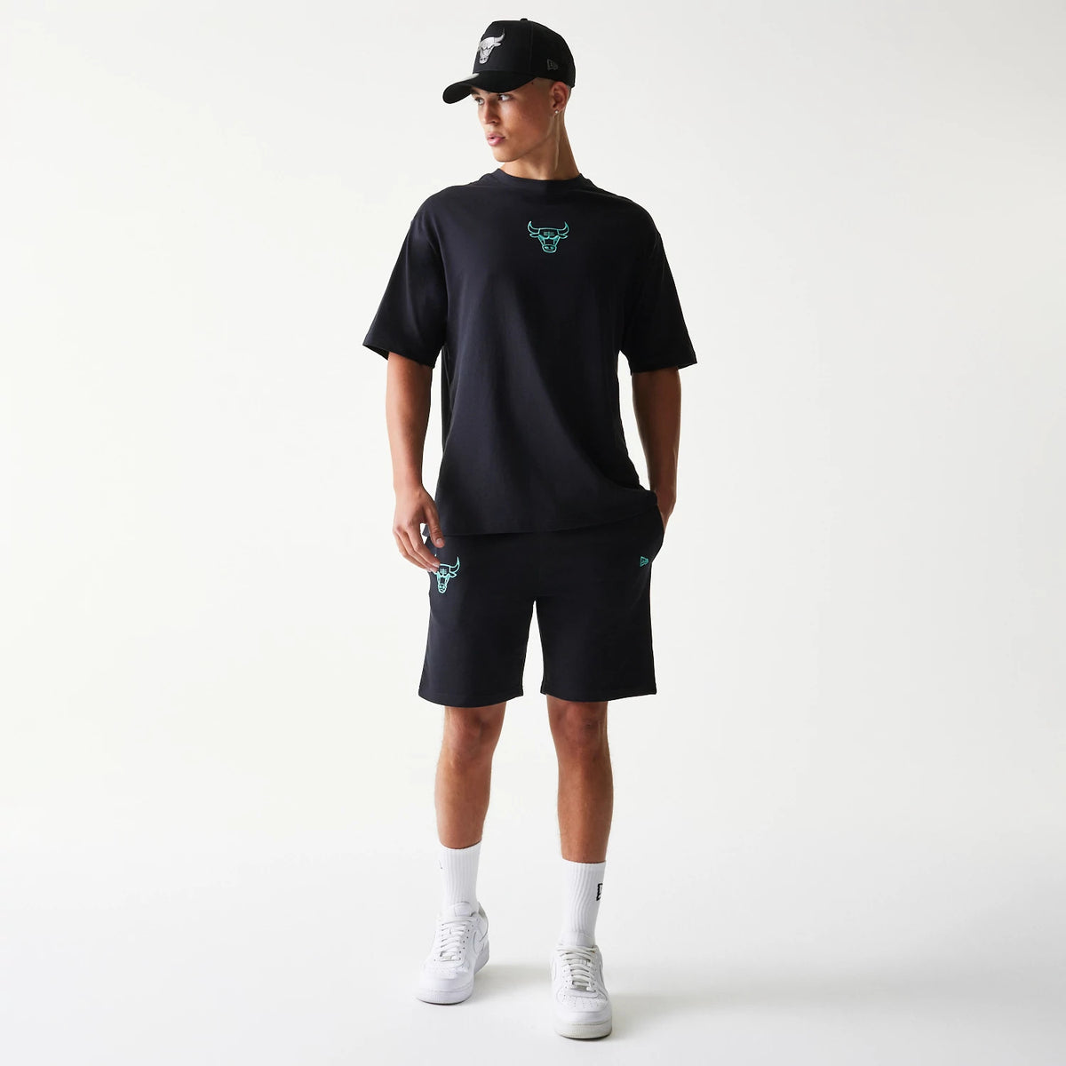 New Era Chicago Bulls NBA League Essential Black Shorts, on-model front view showing the teal embroidered Bull logo on the left leg and the black colourway, styled with a matching black t-shirt with a teal logo on the chest and a black cap.