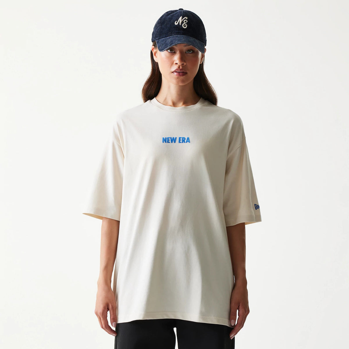New Era Graphic Light Beige Oversized T-Shirt, front view on-model showing the blue printed New Era logo centrally placed on the chest, short sleeves, and a small embroidered New Era logo on the left sleeve.