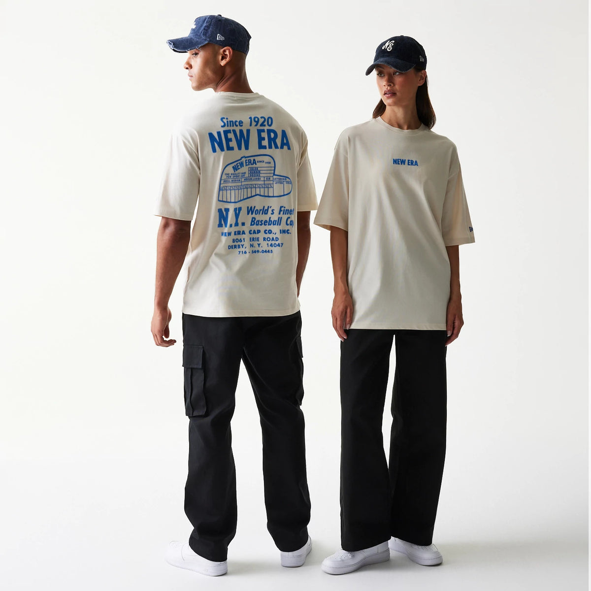 New Era Graphic Light Beige Oversized T-Shirt, back and front view on models with dark caps, showing a large blue graphic and text on the back and a small blue New Era logo embroidered on the chest.
