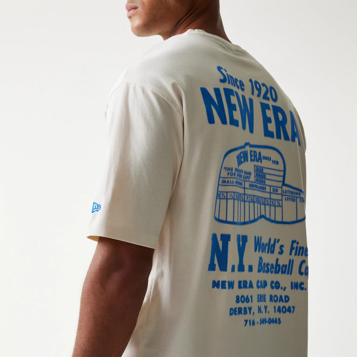 New Era Graphic Light Beige Oversized T-Shirt, back view on a model featuring blue vintage-style graphics and text including New Era across the shoulder blades and the embroidered New Era logo on the sleeve.
