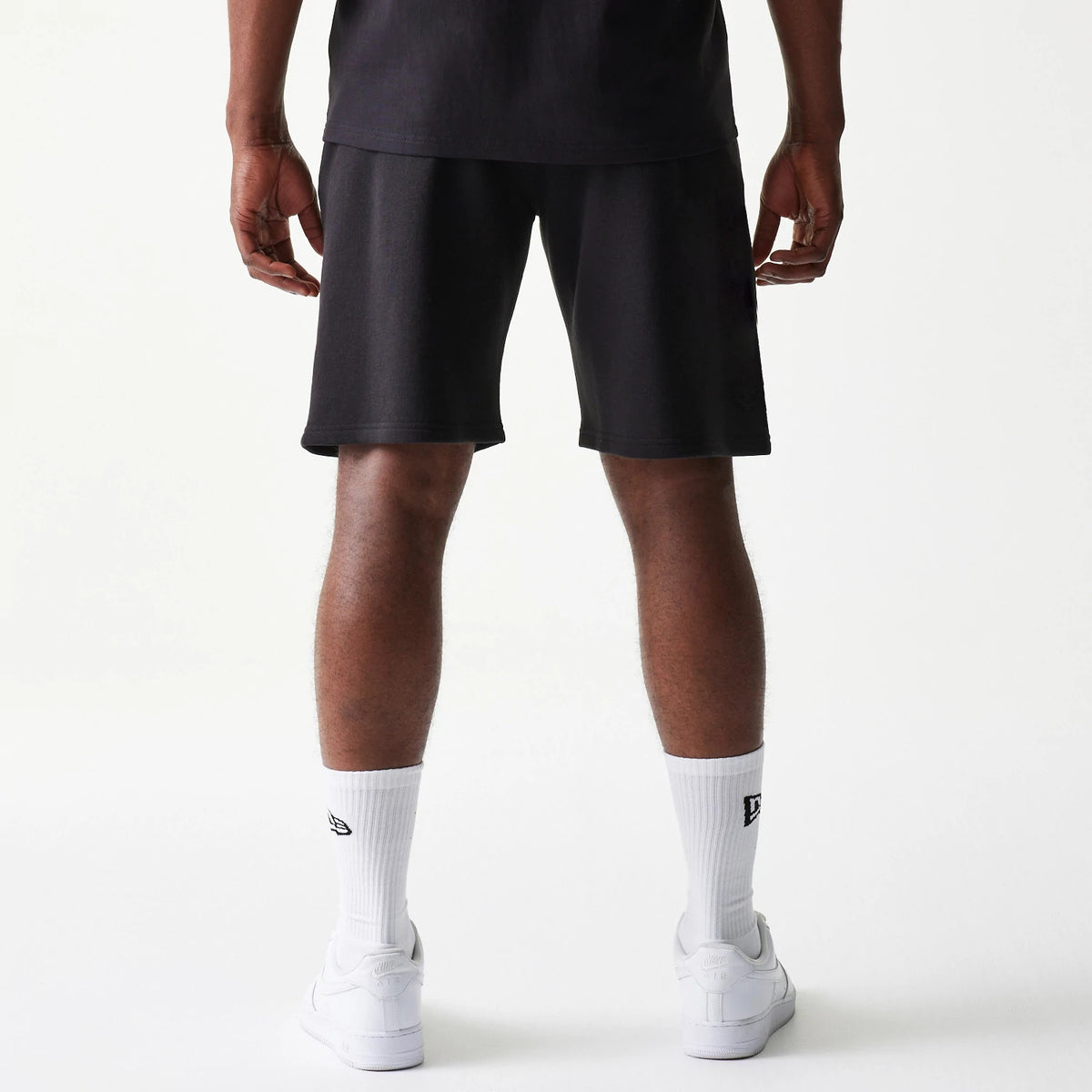 New Era Chicago Bulls NBA Black Shorts, back view showing the plain black fabric on-model, reaching above the knee with a loose fit, paired with white New Era logo socks and trainers for a casual look.