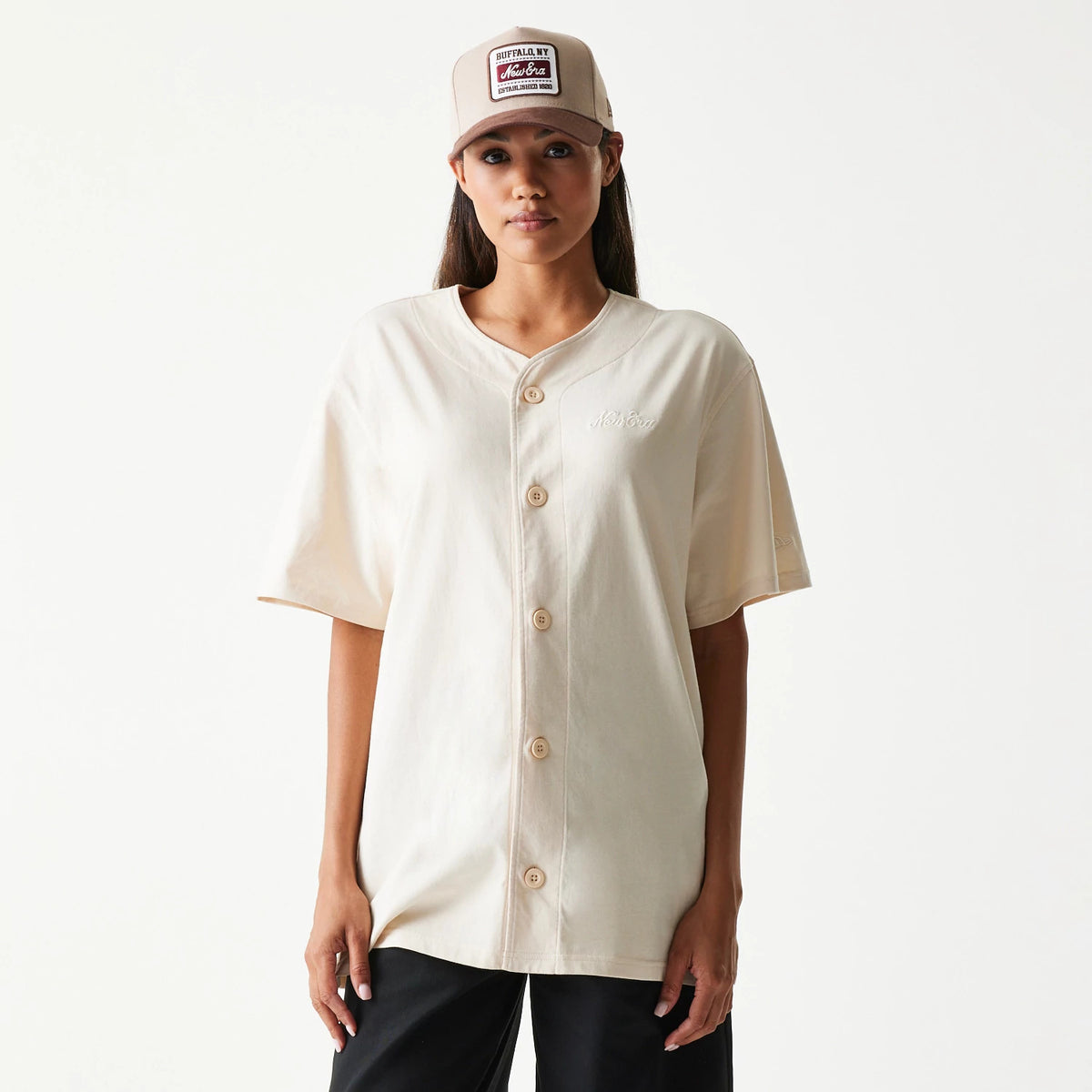 New Era Script Light Beige T-Shirt, front view on a model styled with black trousers and a beige cap, featuring a button-down front and a embroidered New Era logo on the left chest.