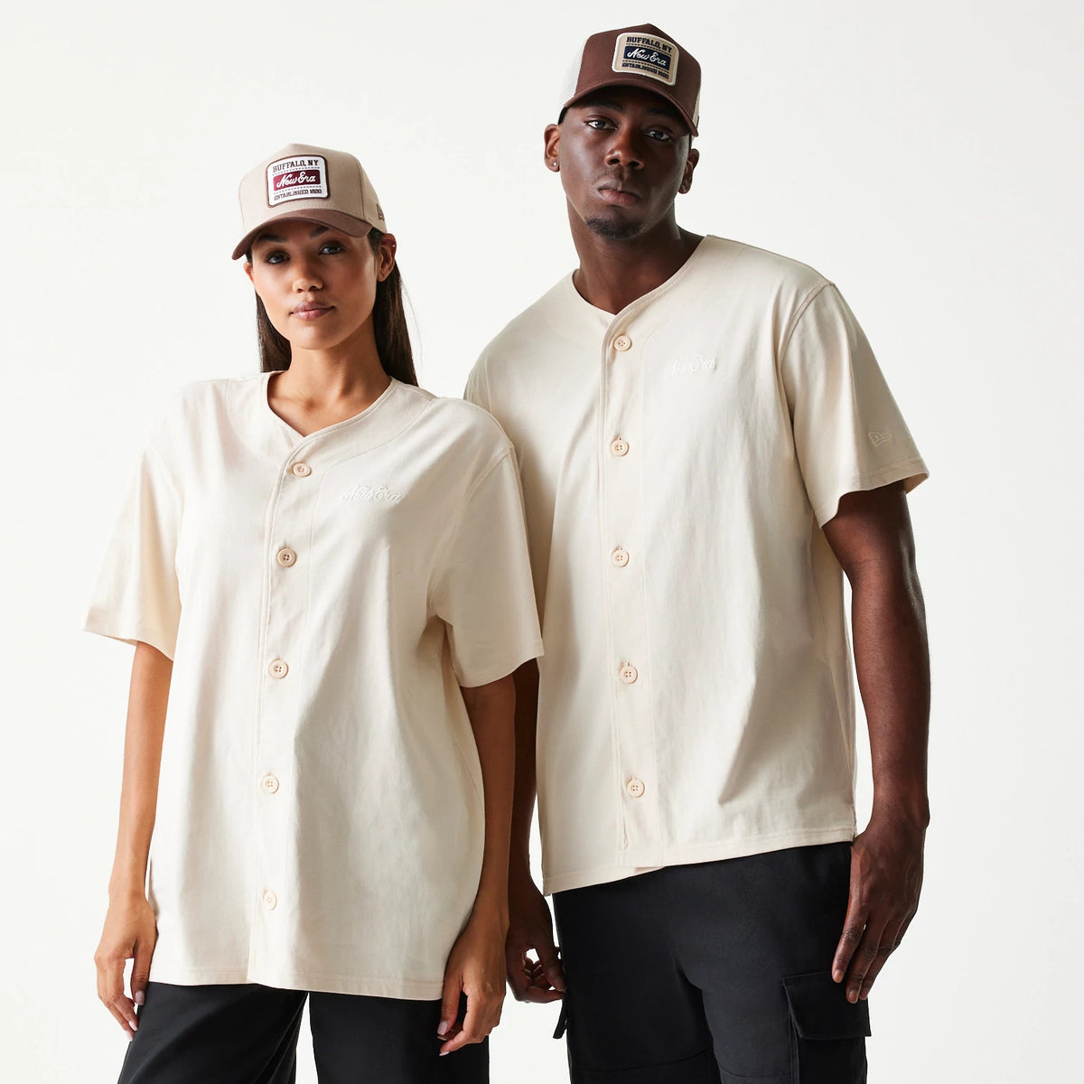 New Era Script Light Beige T-Shirt, front view on a model with a matching cap showing the button-up design with large buttons, embroidered script logo on the chest, and short sleeves, styled with casual black trousers.