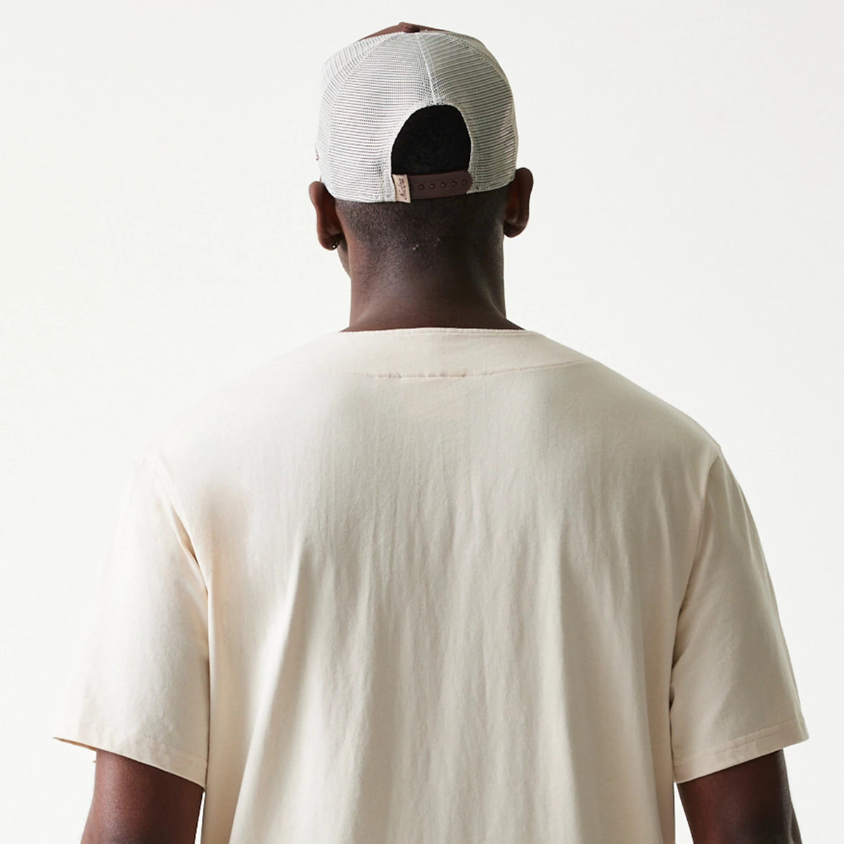 New Era Script Light Beige T-Shirt, back view on a model wearing a mesh cap, showing the plain light beige fabric with short sleeves and a simple round neckline, highlighting the clean and understated design.