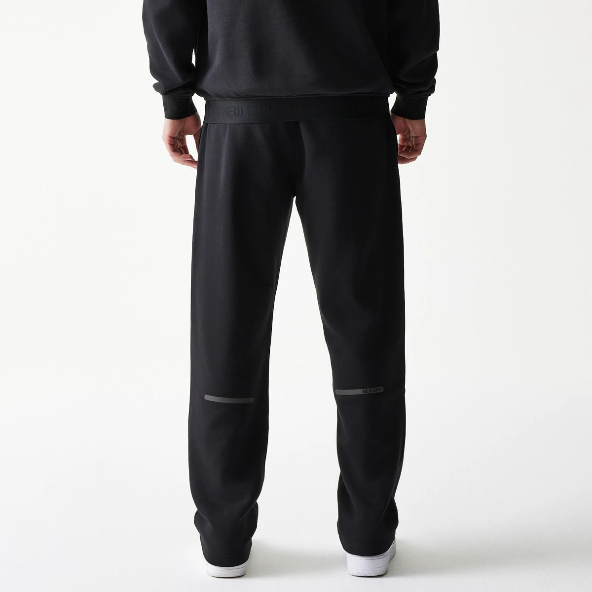 New Era Tech Black on Black Straight Leg Joggers, back view on a model showing the elastic waistband with branding, straight leg fit with grey horizontal accents above the knees, and white trainers.