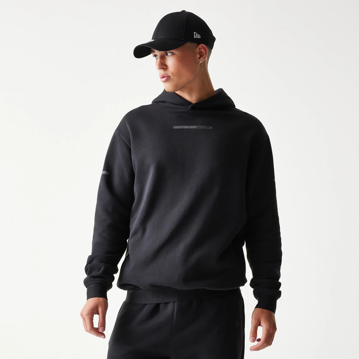 New Era Tech Strip Black Oversized Pullover Hoodie, front view on-model showing the black design with a strip logo across the chest and plain long sleeves, styled with matching black joggers and a black cap.