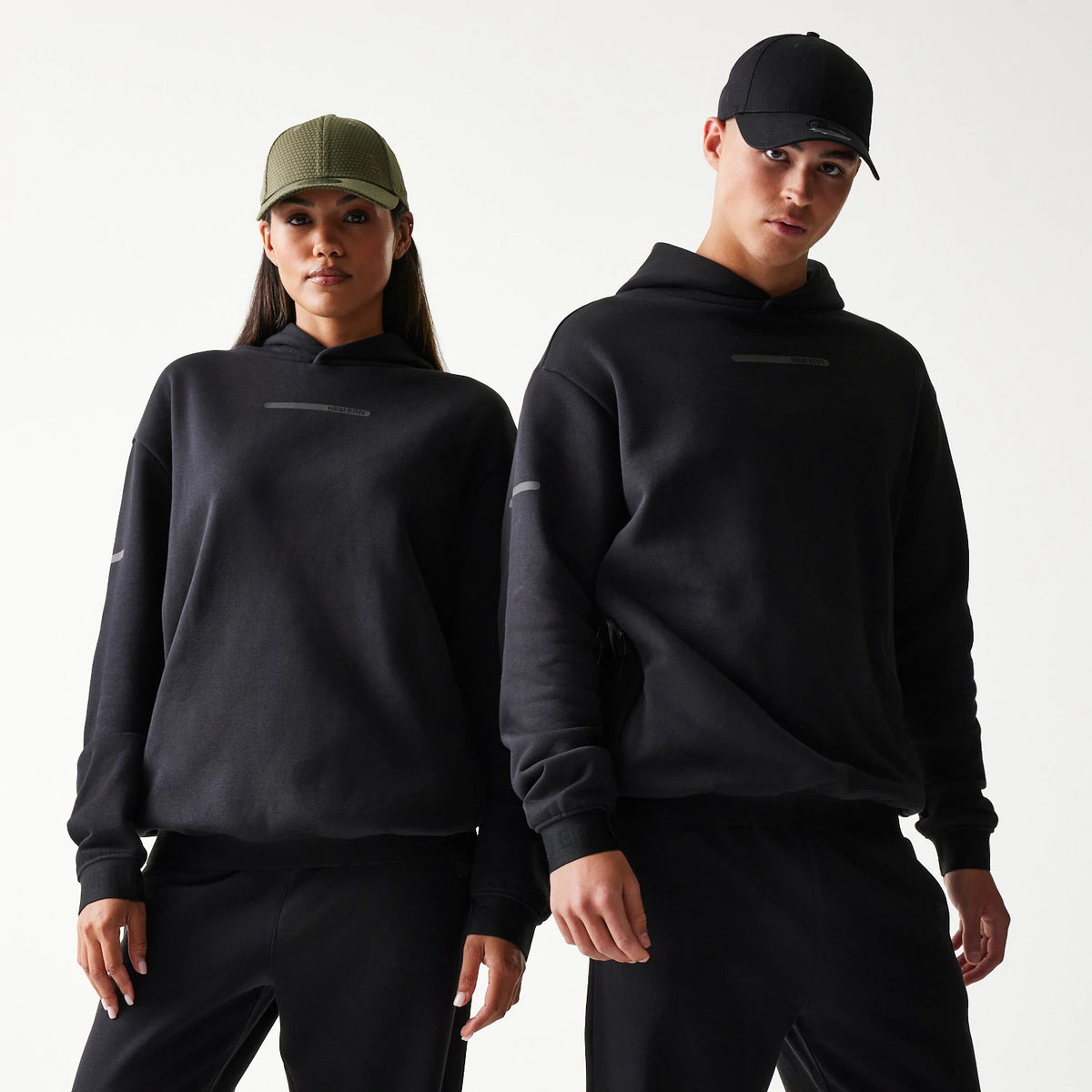 New Era Tech Strip Black Oversized Pullover Hoodie, front view on-model featuring a tech strip design on the chest, long sleeves with faint on the left arm, styled with matching black trousers and caps.