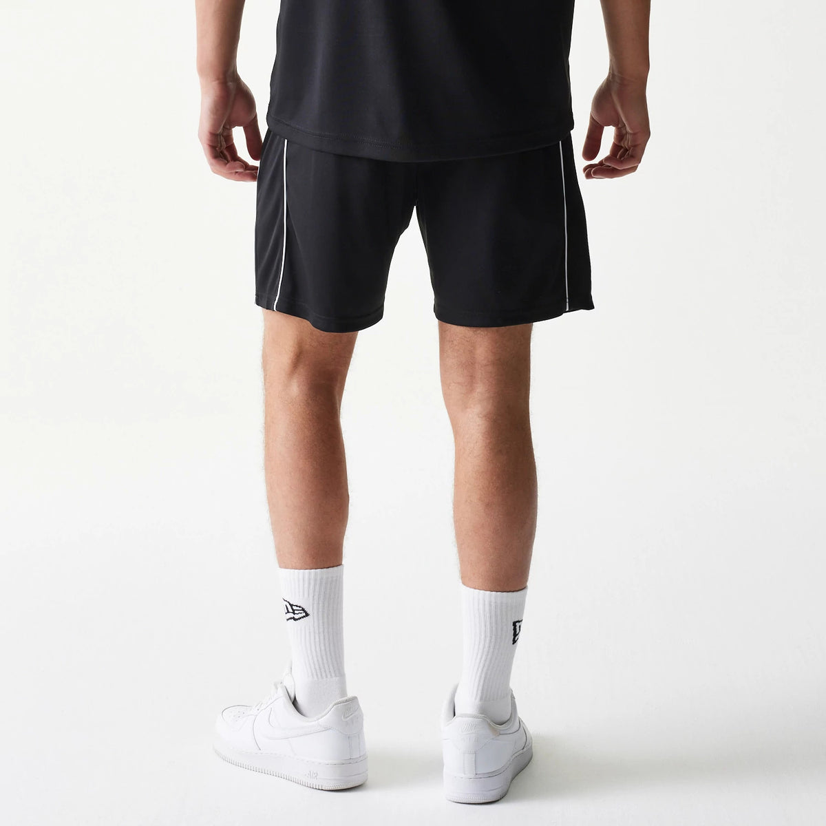 New Era Football Black Shorts, back view on a model featuring black shorts with white piping along the sides, paired with white socks showcasing the embroidered New Era logo above clean white trainers.