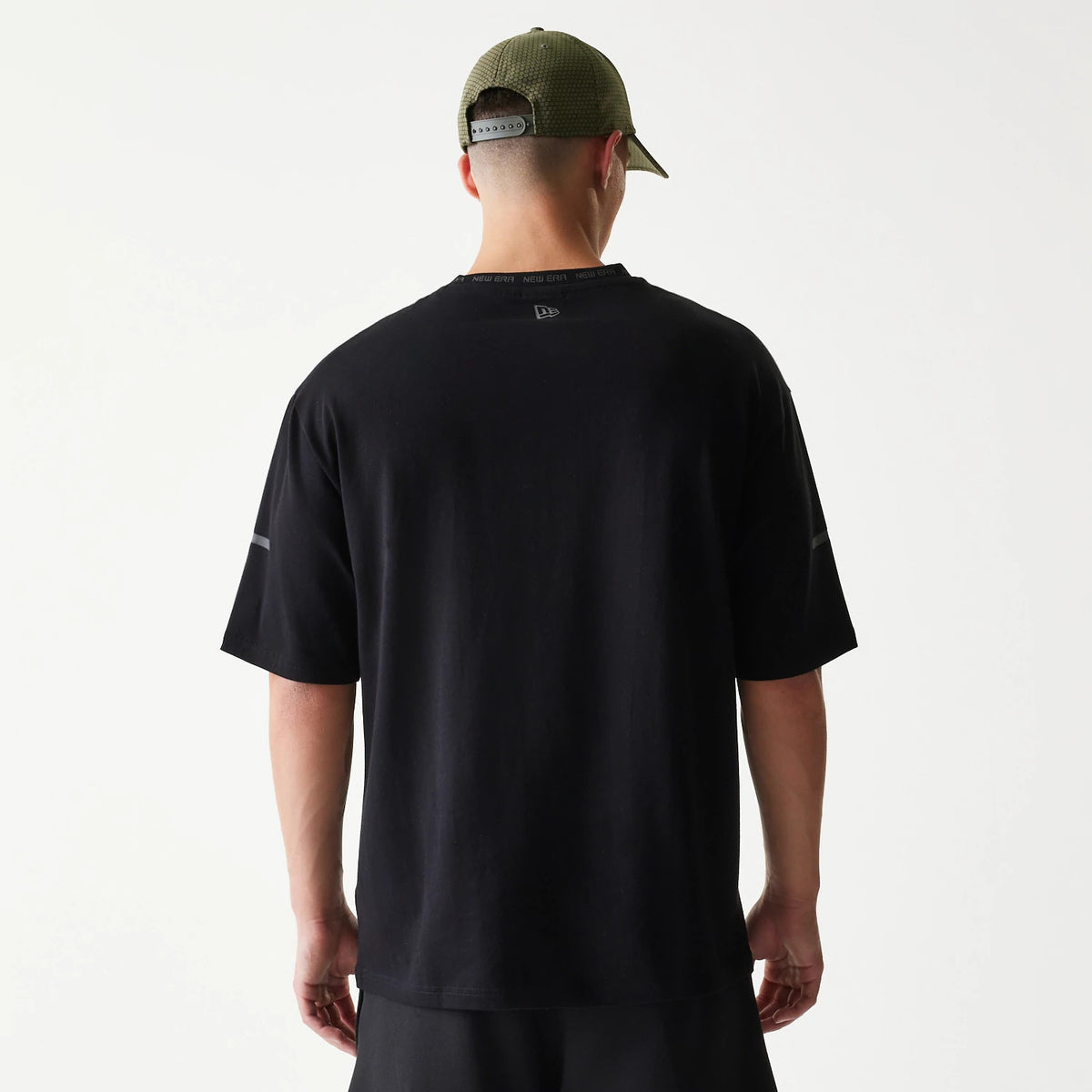 New Era Tech Black Oversized T-Shirt, back view on-model featuring a grey embroidered New Era logo at the upper centre of the back, with a loose fit and short sleeves, styled with a green cap.