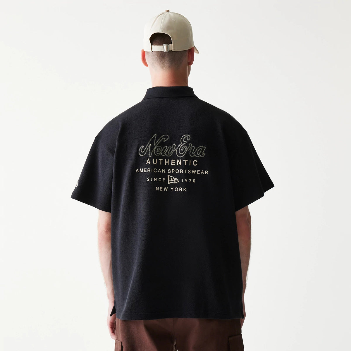 New Era Graphic Black Polo T-Shirt, back view on a model pairing it with brown shorts and a beige cap, featuring embroidered New Era Authentic lettering and American Sportswear text centred on the upper back.