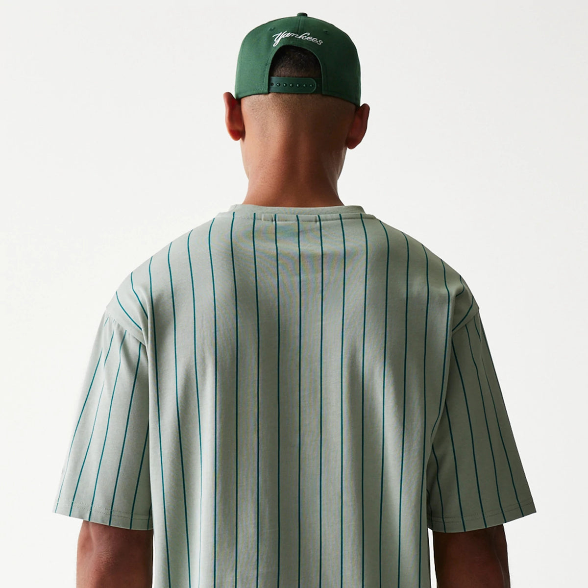 New Era Pinstripe Pastel Green Oversized T-Shirt, back view on a model showing vertical pinstripes running down from the shoulders to the hem, paired with a green cap featuring an embroidered script logo on the back.