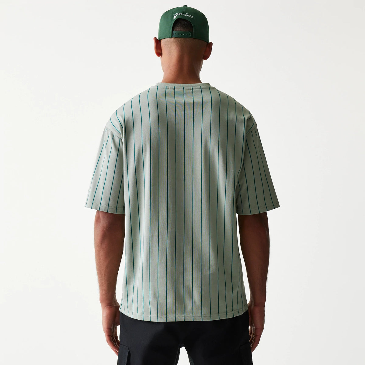 New Era Pinstripe Pastel Green Oversized T-Shirt, back view on-model displaying vertical pinstripes running down the pale green fabric, with short sleeves and a loose fit that drapes over dark trousers, paired with a green cap.