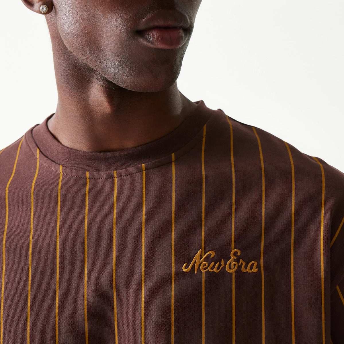 New Era Pinstripe Brown Oversized T-Shirt, front view on-model highlighting the vertical yellow pinstripes on a brown background and an embroidered New Era logo in yellow on the left chest.