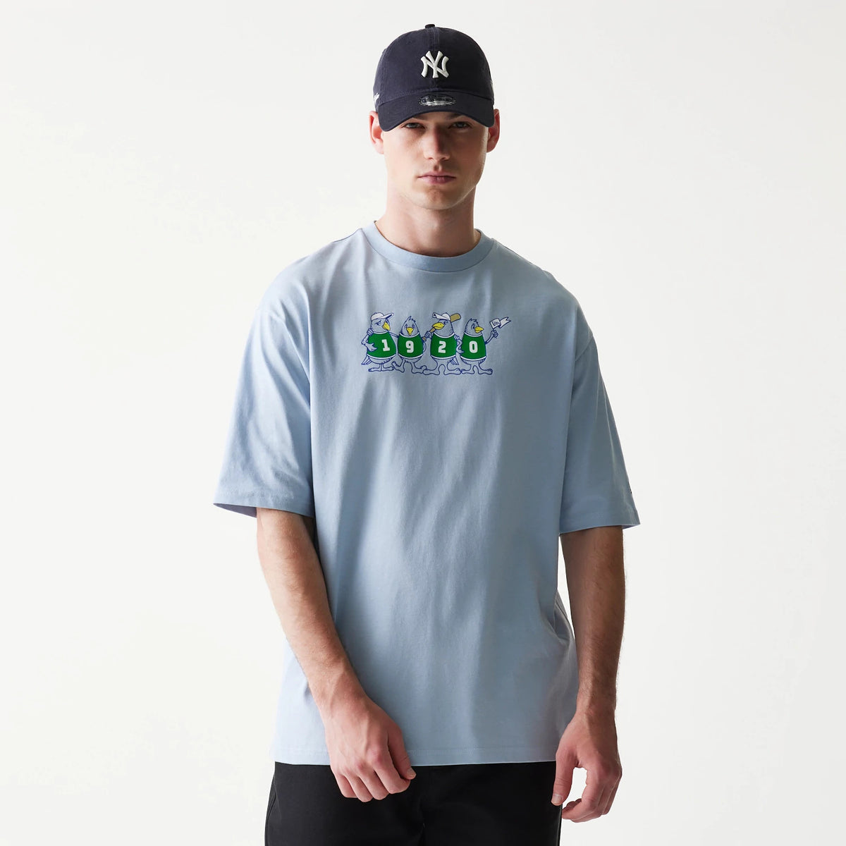 New Era Baseball Birds Character Graphic Blue Oversized T-Shirt, front view on a model showing an animated bird design with the numbers 1920 across the chest, paired with a dark cap featuring an embroidered logo.