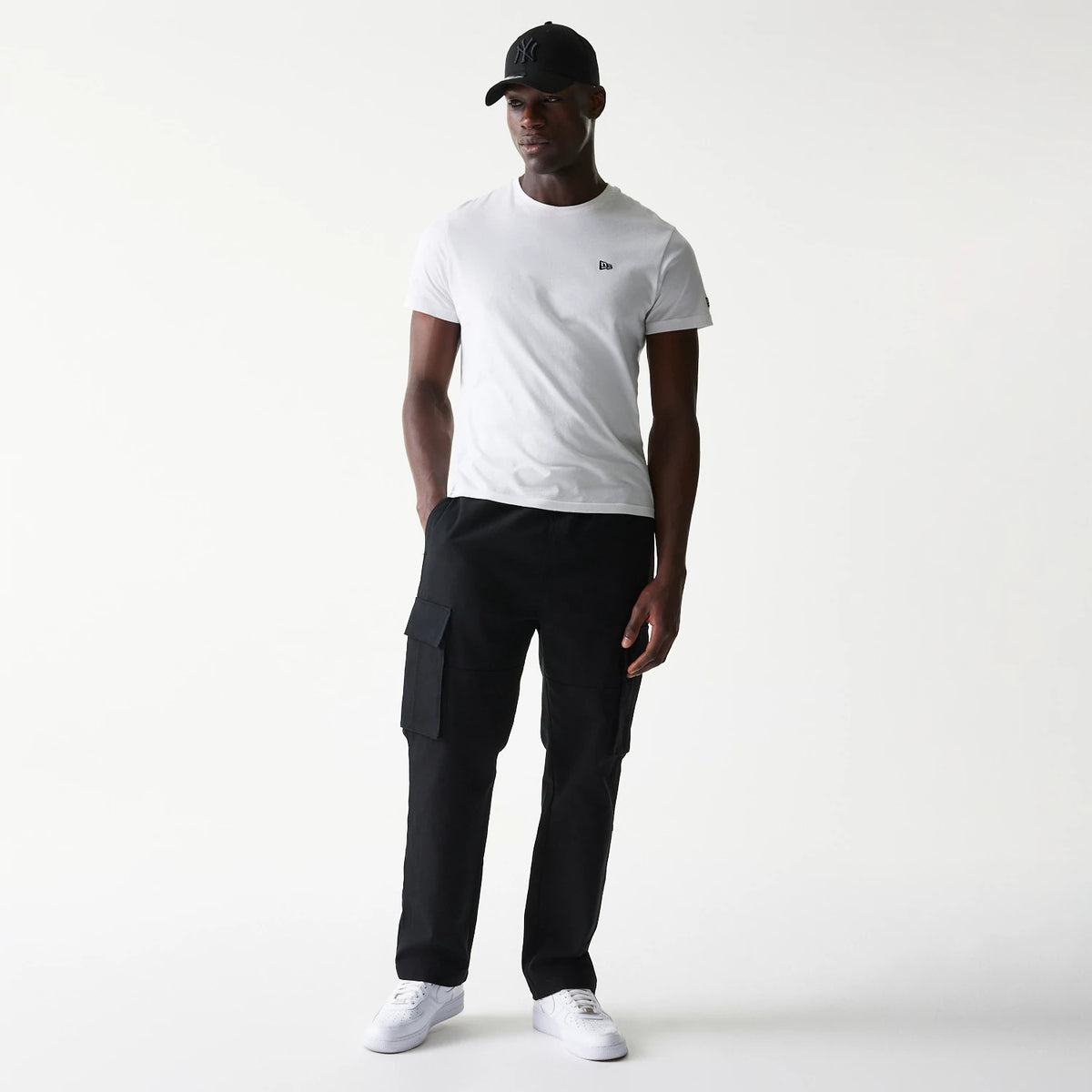 New Era Black Straight Leg Cargo Joggers, front view on a model wearing a white T-shirt and black cap, showing the joggers' side cargo pockets and relaxed fit with hands in pockets.