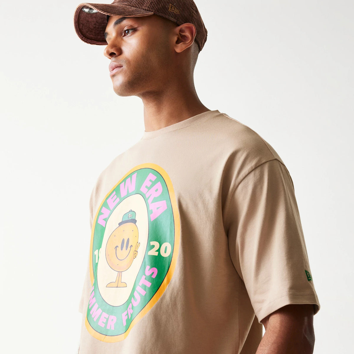 New Era Orange Fruit Graphic Light Beige Oversized T-Shirt, front three-quarter view on-model showing the large colourful fruit graphic with New Era branding on the chest and a small green embroidered New Era logo on the left sleeve.