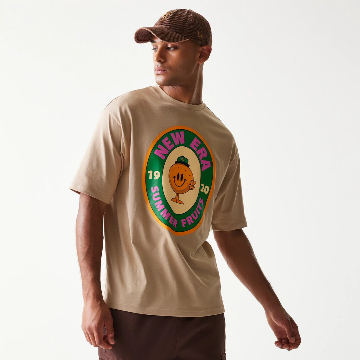 New Era Orange Fruit Graphic Light Beige Oversized T-Shirt, front view on-model showing a large fruit graphic with New Era Summer Fruits lettering and a cheerful orange character centred on the light beige fabric.