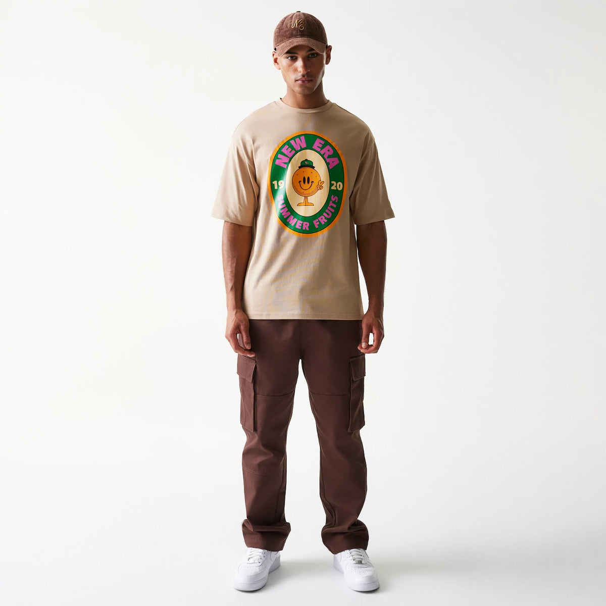 New Era Orange Fruit Graphic Light Beige Oversized T-Shirt, front view on-model featuring a colourful graphic of a smiling fruit wearing a green cap with New Era branding, displayed centrally on the light beige fabric.