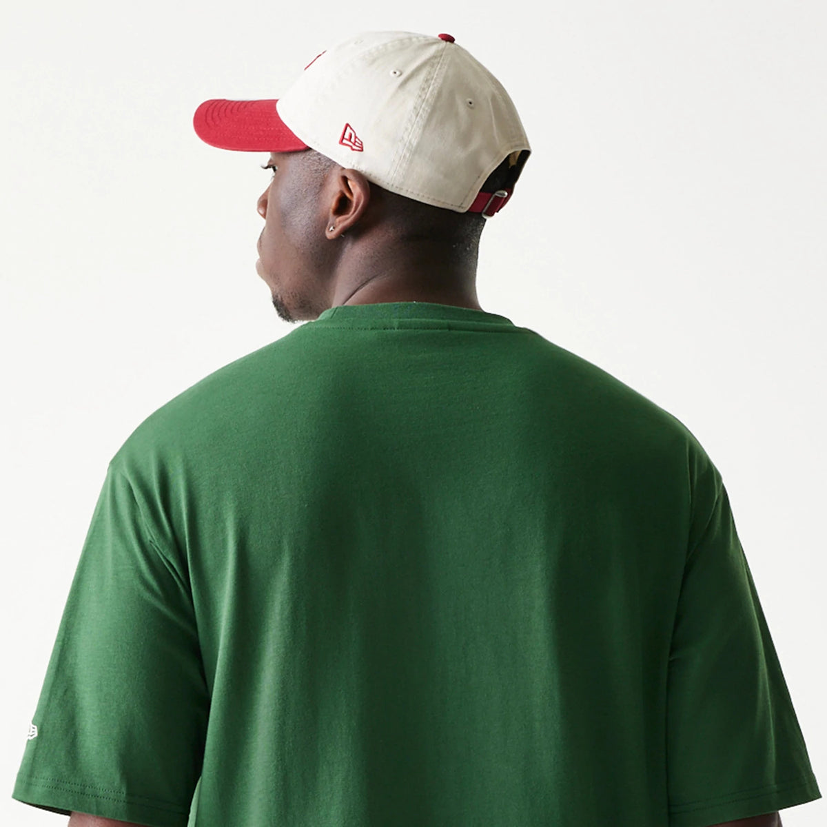 New Era Pear Fruit Graphic Dark Green Oversized T-Shirt, back view on a model featuring a plain dark green back with short sleeves, paired with a cream cap showing the embroidered New Era logo on the side and a red visor.