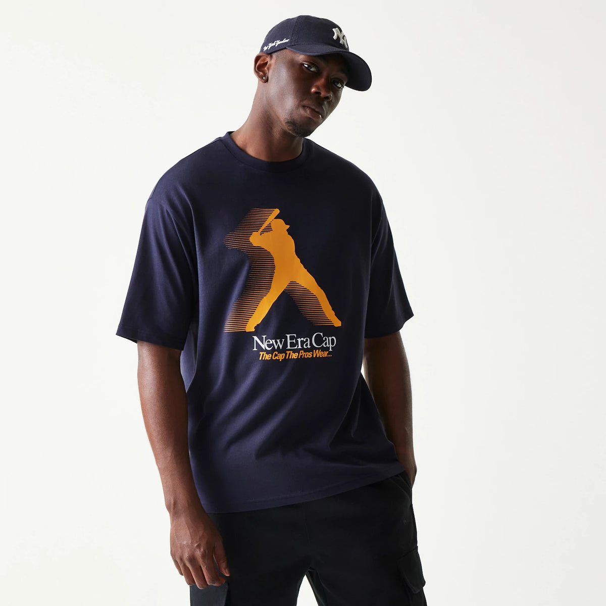 New Era Baseball Player Graphic Navy Oversized T-Shirt, front view on a model with a navy cap, showcasing a orange baseball player graphic and white New Era Cap lettering with the slogan The Cap The Pros Wear.