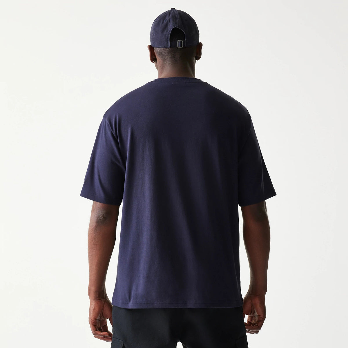New Era Baseball Player Graphic Navy Oversized T-Shirt, back view on a model featuring a plain navy design with short sleeves and a relaxed fit, paired with a navy cap showing a small back adjustment strap.