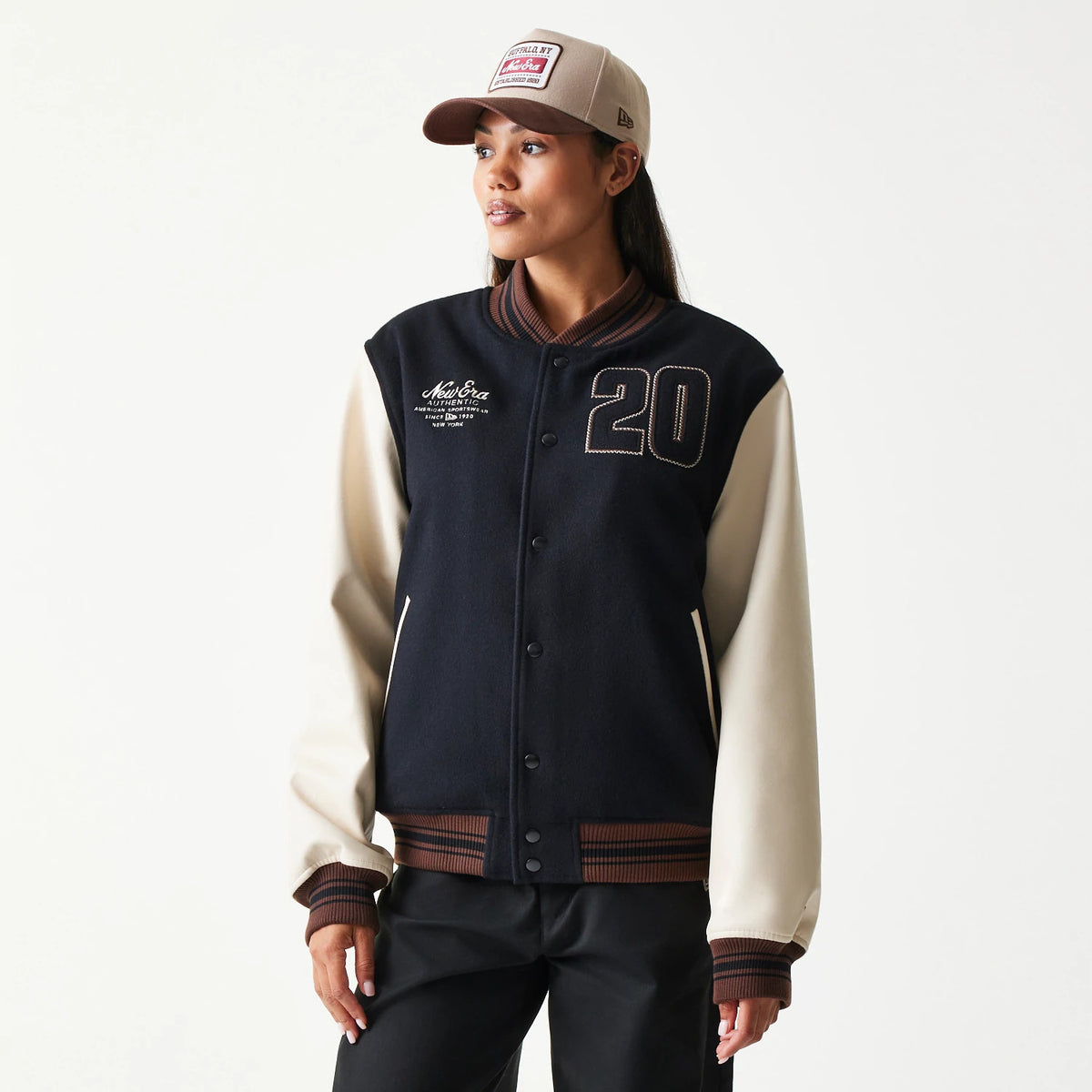 New Era Varsity Black Jacket, front view on-model showing cream sleeves, central snap closure, embroidered 20 on the right chest, New Era Authentic script on the left, and striped brown and black collar and cuffs.