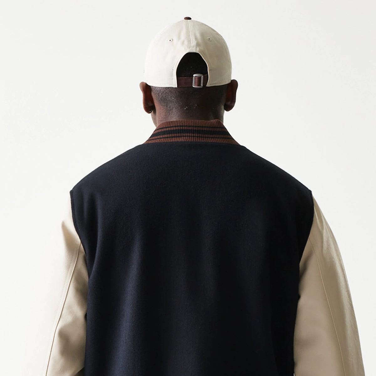 New Era Varsity Black Jacket, back view on-model highlighting the contrasting cream sleeves with a black body, a ribbed collar with brown stripes, and an overall classic varsity silhouette.