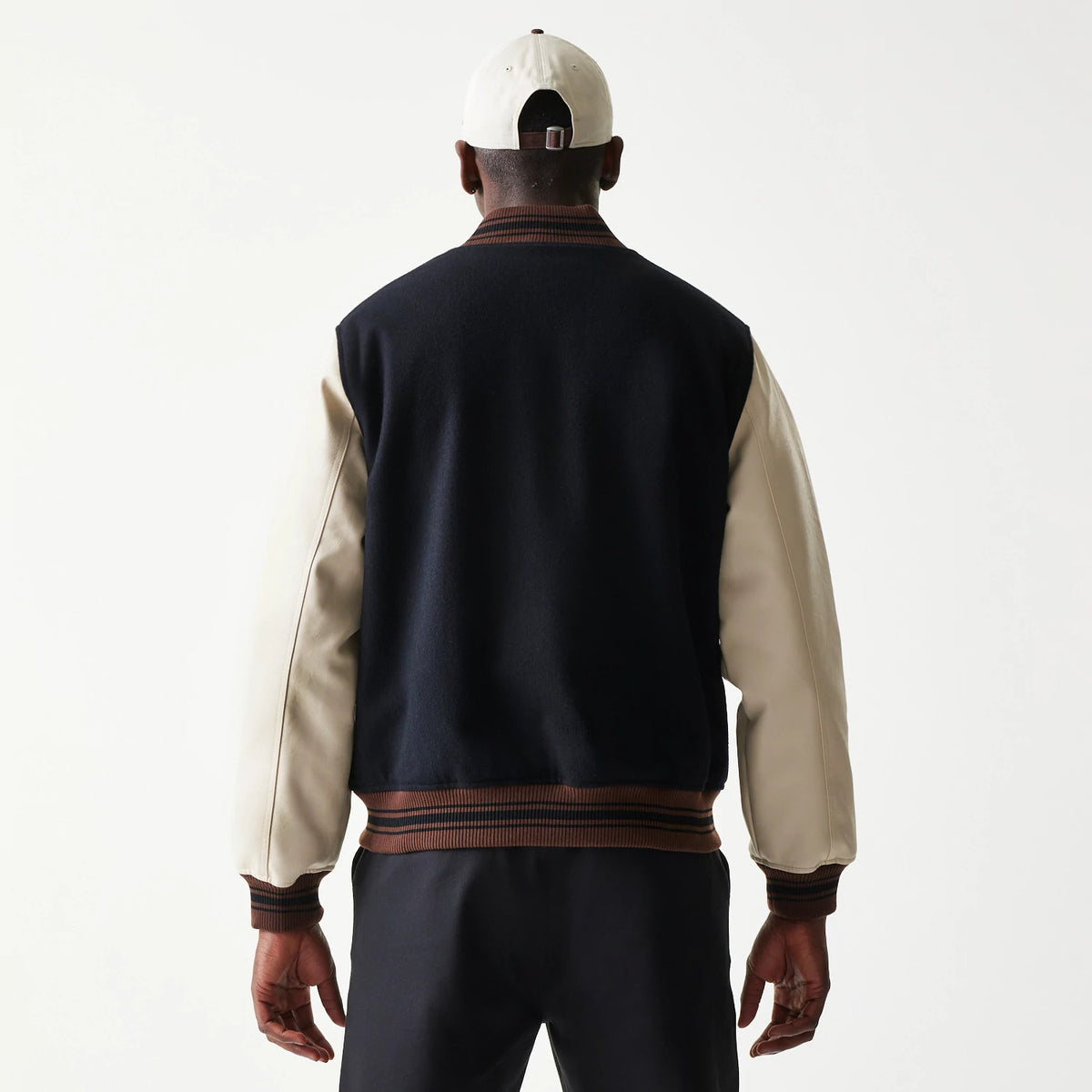 New Era Varsity Black Jacket, back view on a model with a contrasting cap, highlighting the black body and cream sleeves with brown ribbed cuffs and hem, creating a classic varsity style.