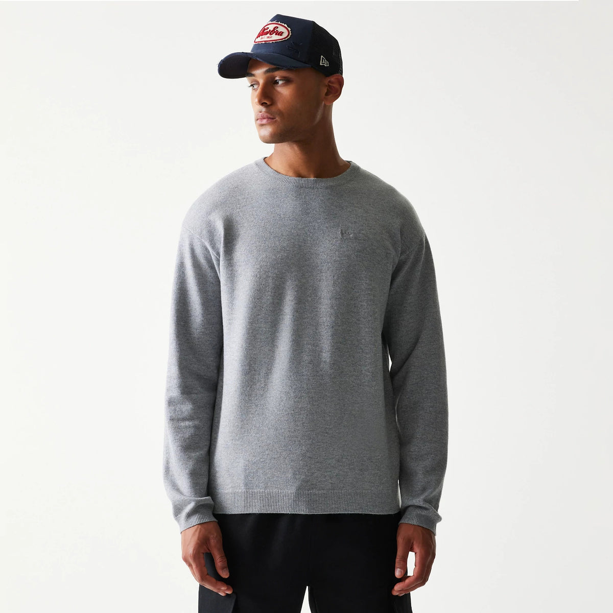 New Era Essential Flag Grey Knitted Sweater, front view on a model, showcasing a simple crew neck design with long sleeves and ribbed cuffs and hem, paired with a dark cap featuring an embroidered front logo and New Era flag logo on the side.