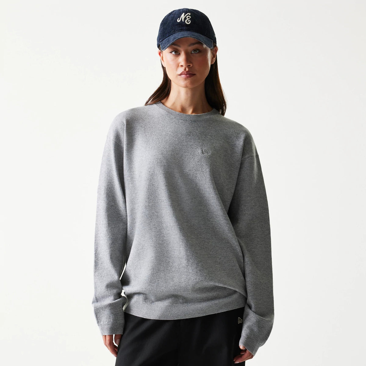 New Era Essential Flag Grey Knitted Sweater, front view on a model showing the embroidered logo on the chest and ribbed cuffs, paired with a navy cap, highlighting the soft grey knit fabric.