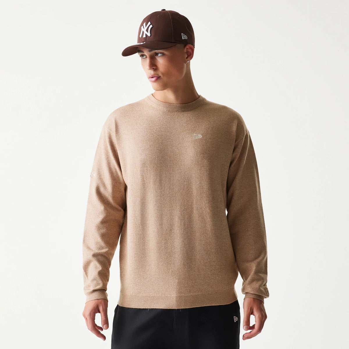 New Era Essential Flag Light Beige Knitted Sweater, front view on a model wearing a brown cap, showing a embroidered New Era flag logo on the left chest and ribbing at the cuffs and hem.
